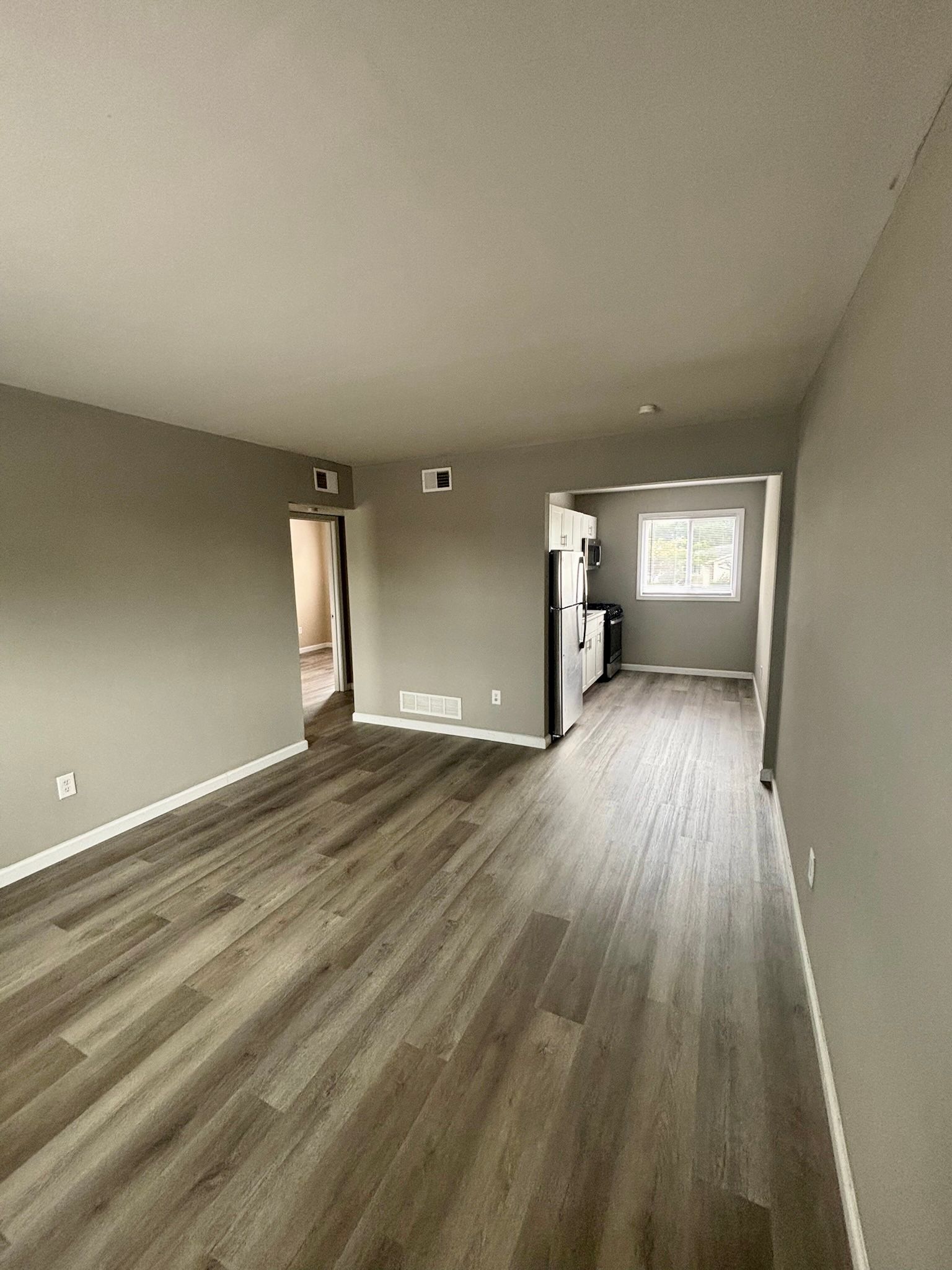 Empty gray room with wood floors and a window, viewed toward a kitchen area.