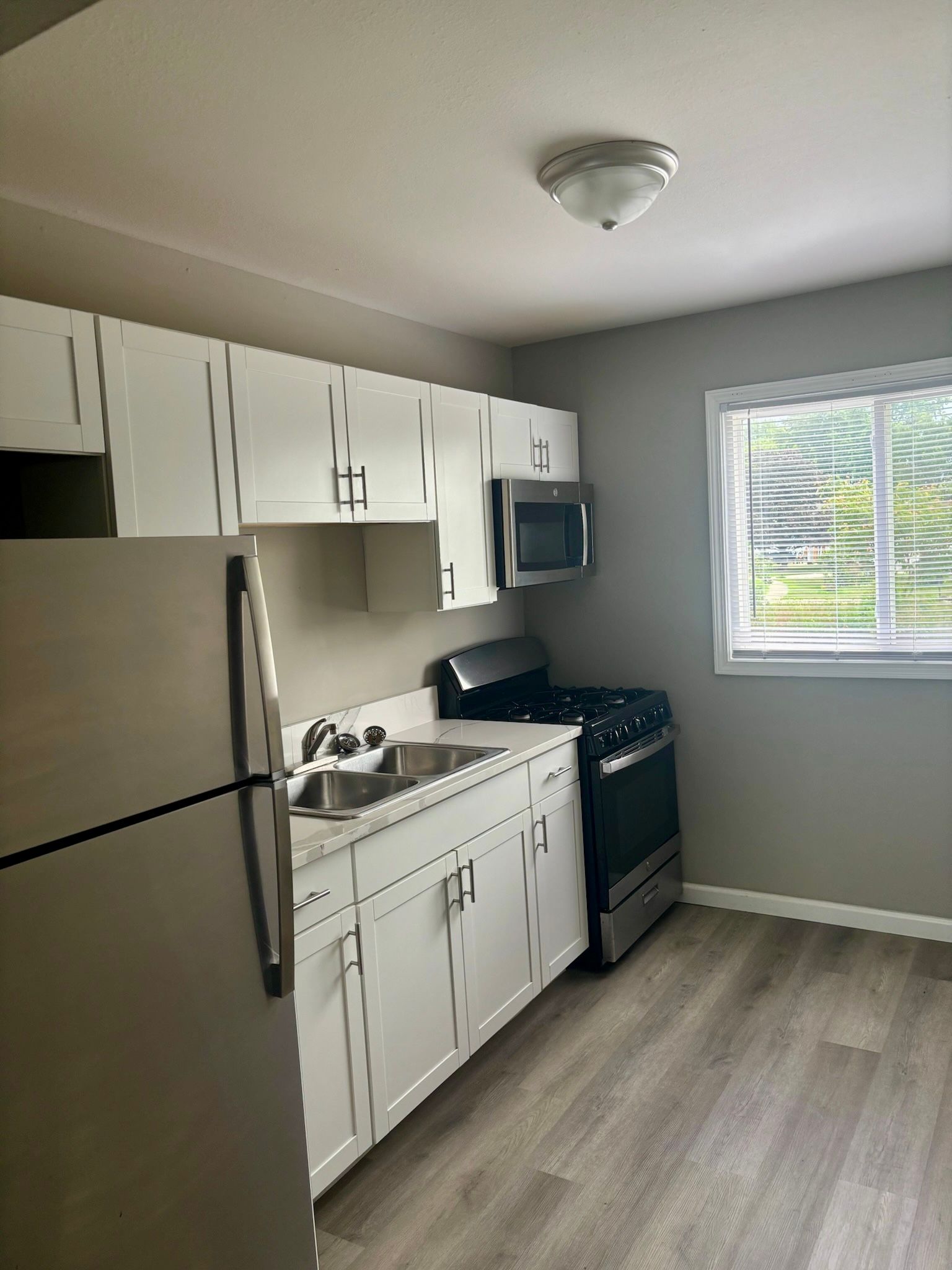 Compact kitchen with white cabinets, stainless fridge, black stove, and bright window on gray walls.