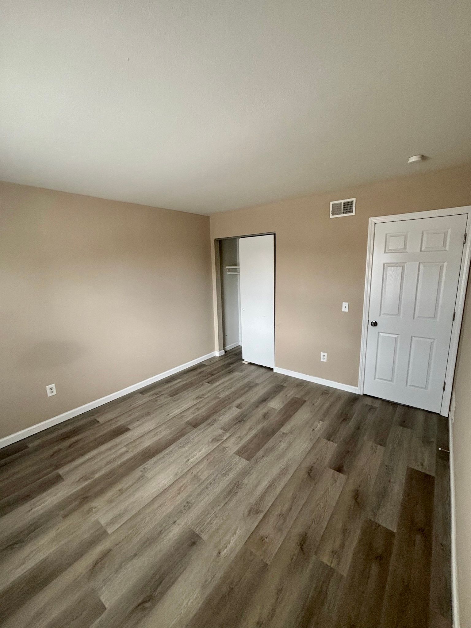 Empty beige bedroom with gray wood flooring, white closet doors, and an open doorway.