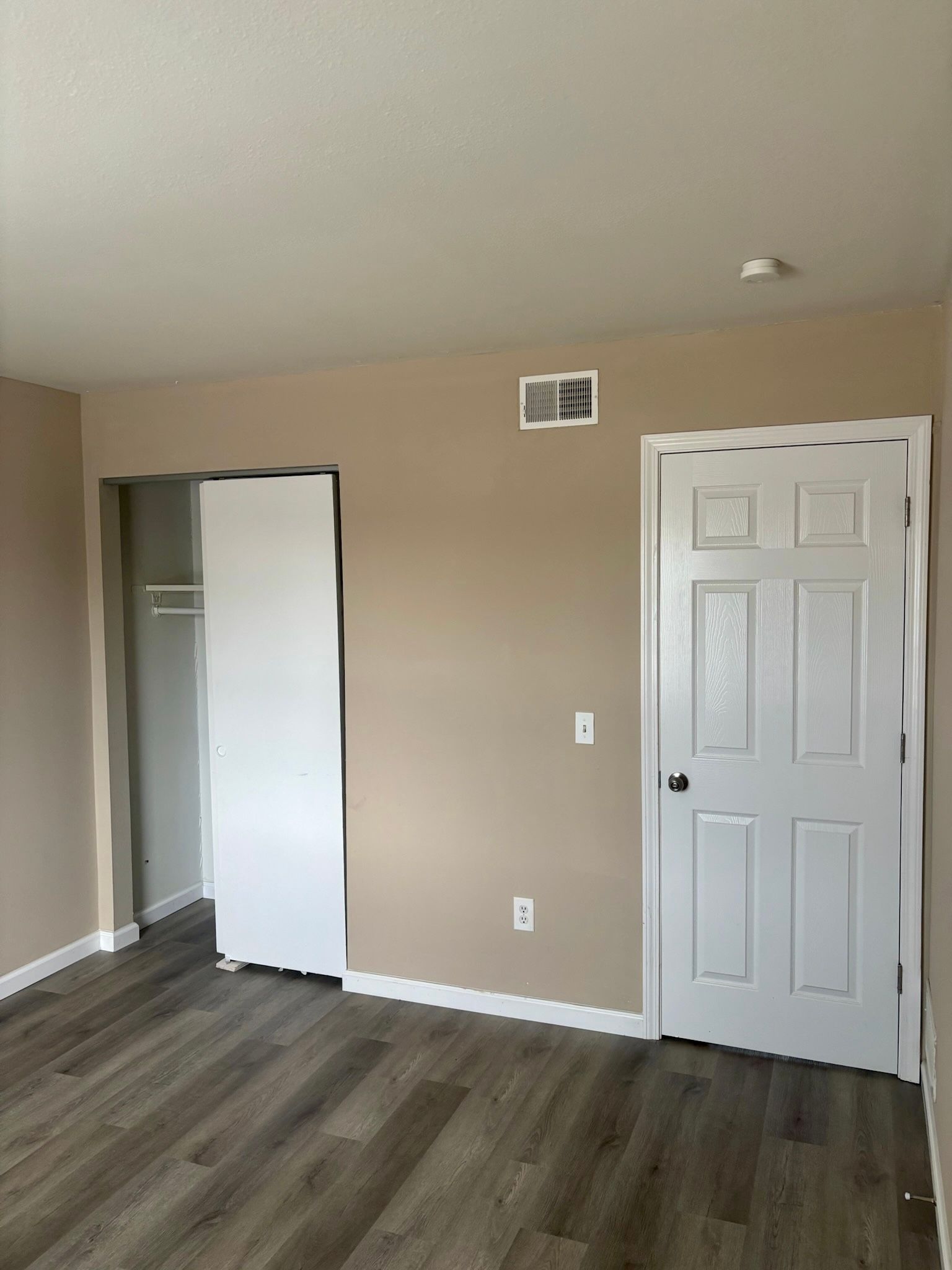 Empty beige bedroom with open closet, white door, and gray wood-look flooring.