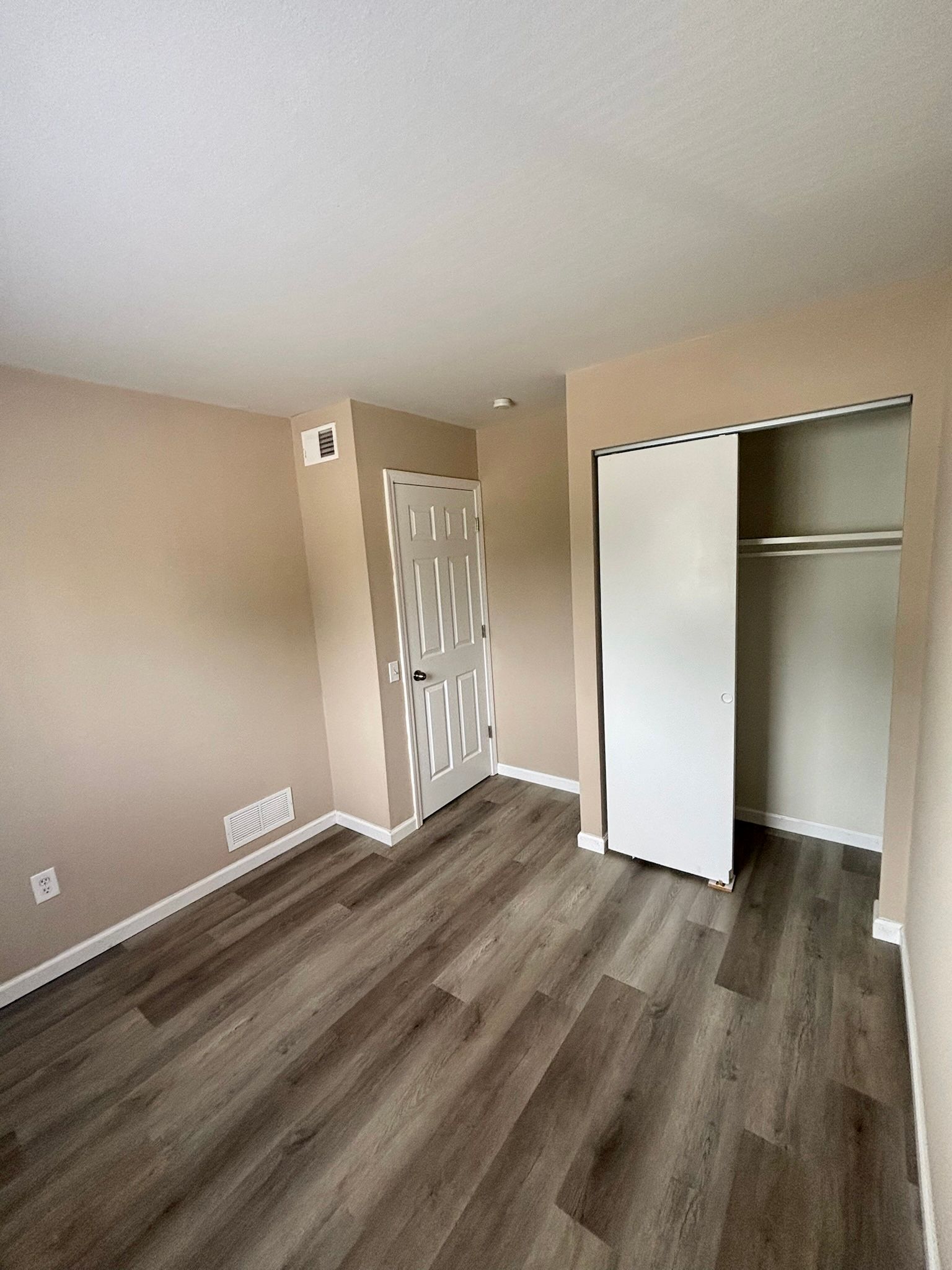 Empty beige bedroom with gray wood flooring, open closet, and doorway to hall