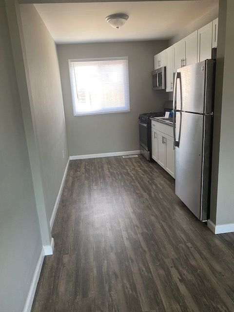 Narrow kitchen with stainless steel fridge, white cabinets, wood-look floor, and a bright window at the end.