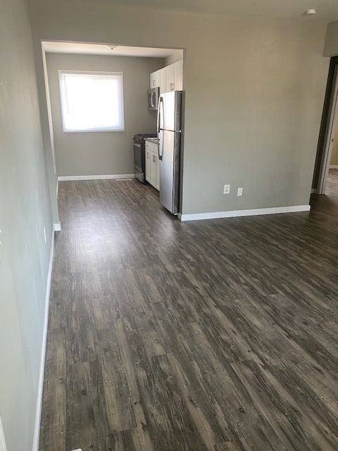 Empty modern kitchen with gray wood flooring, stainless fridge, and a small window in the back.