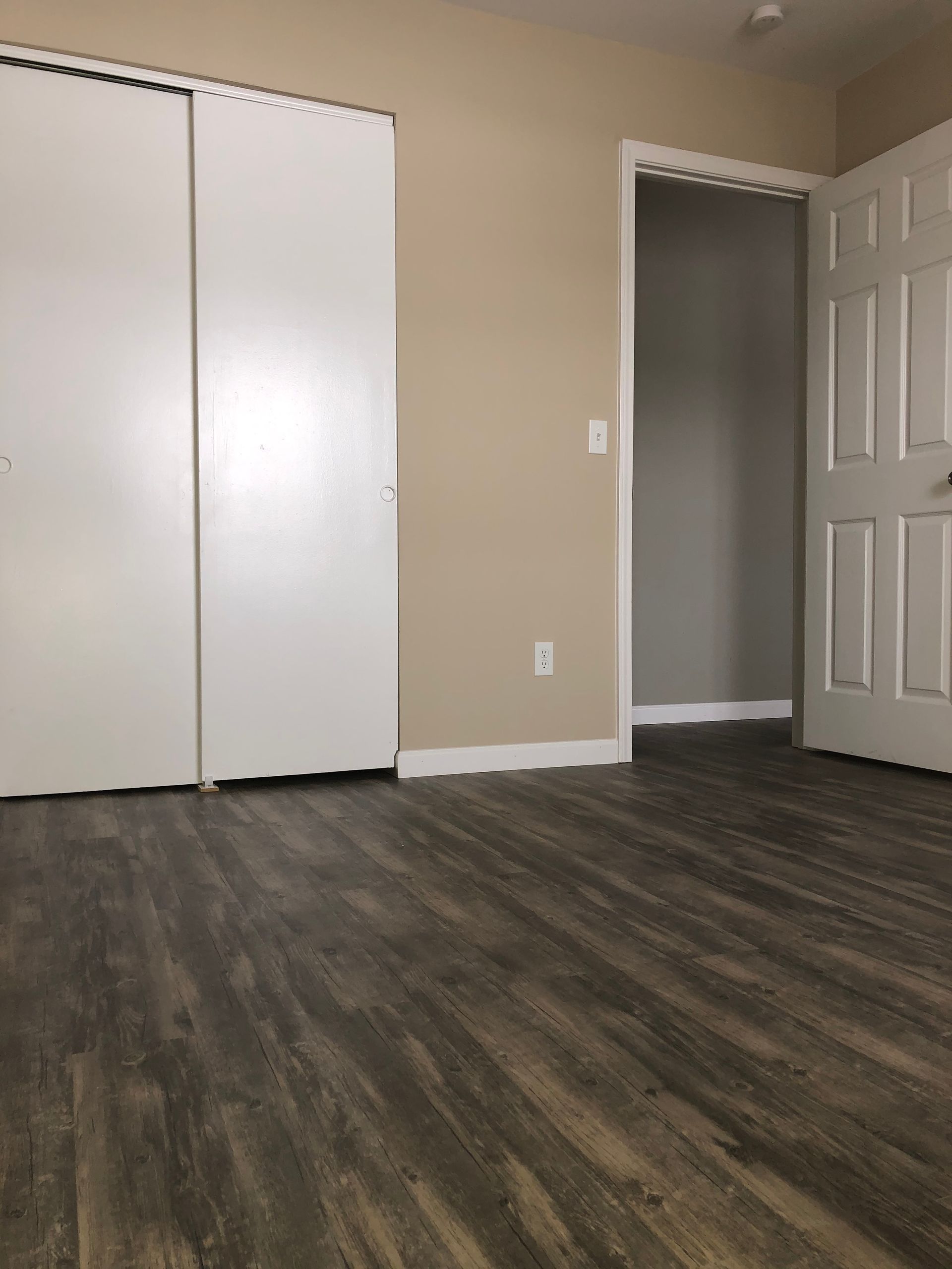 Empty beige bedroom with dark wood floor, white closet doors, and an open white door.
