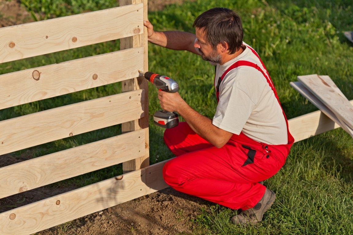 Worker in red overalls drilling a wooden fence panel outdoors.