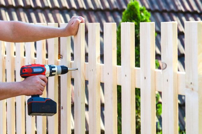 Hand using a power drill to install a white wooden fence panel outdoors