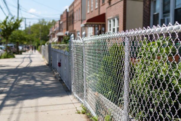 Residential sidewalk beside a chain-link fence and brick rowhouses on a sunny day