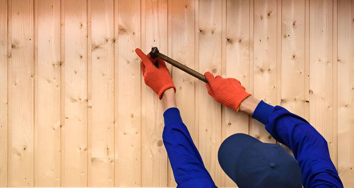 Gloved hands use a hammer and nail on a light wood wall paneling