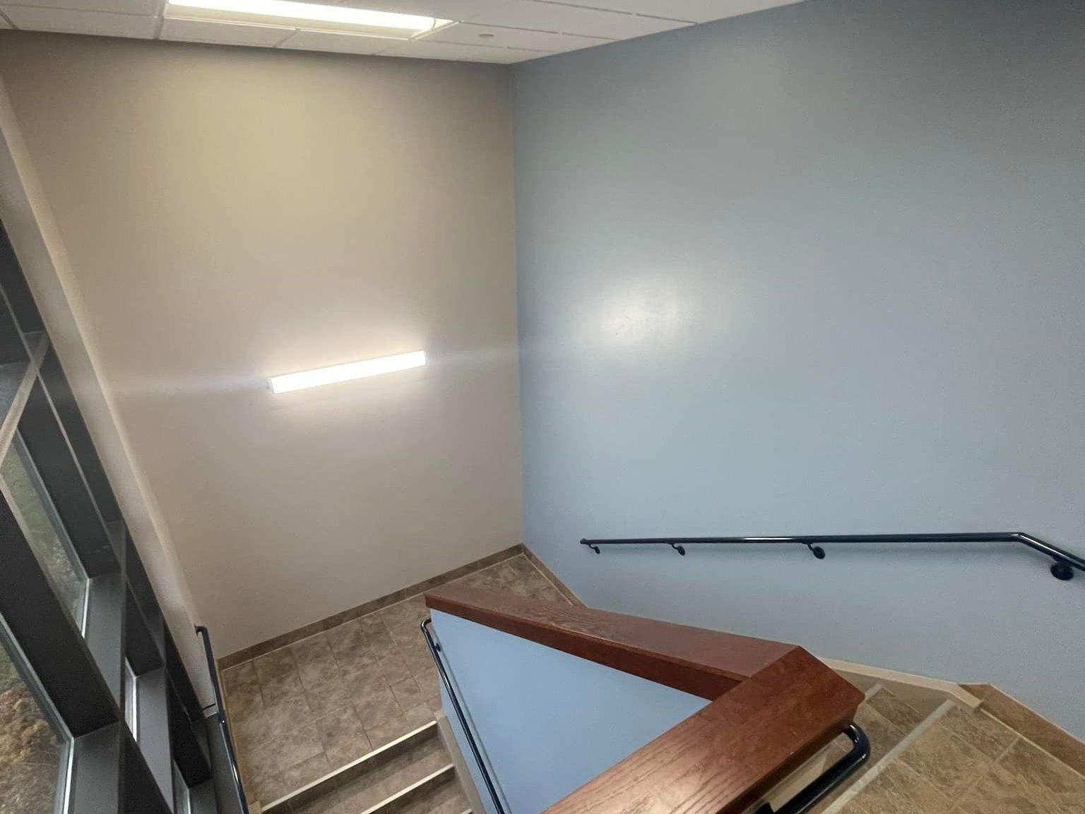 Stairwell with light fixture, gray and blue walls, brown handrail and stairs.