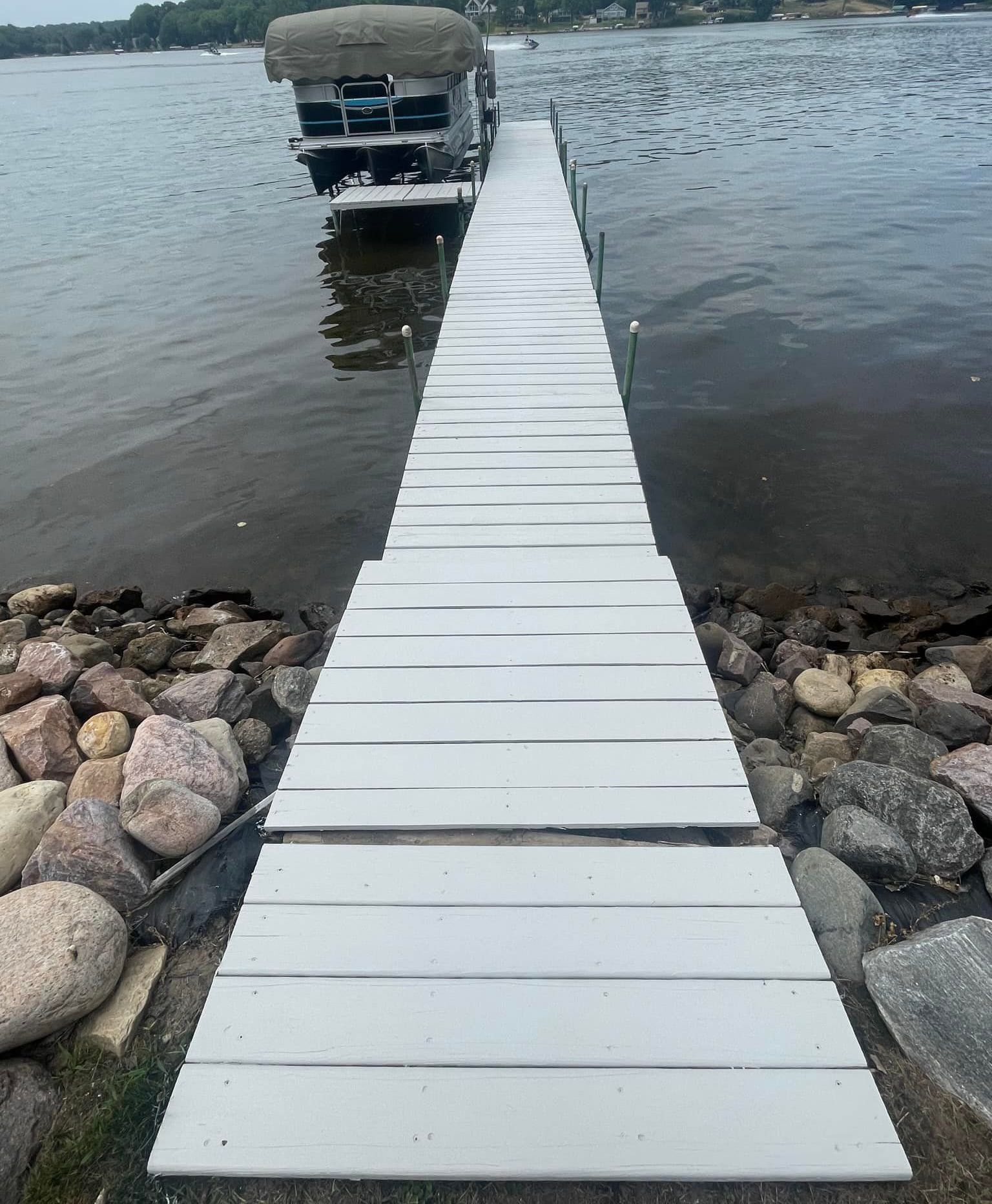 White dock leading to a pontoon boat on a lake. Rocks line the shore.