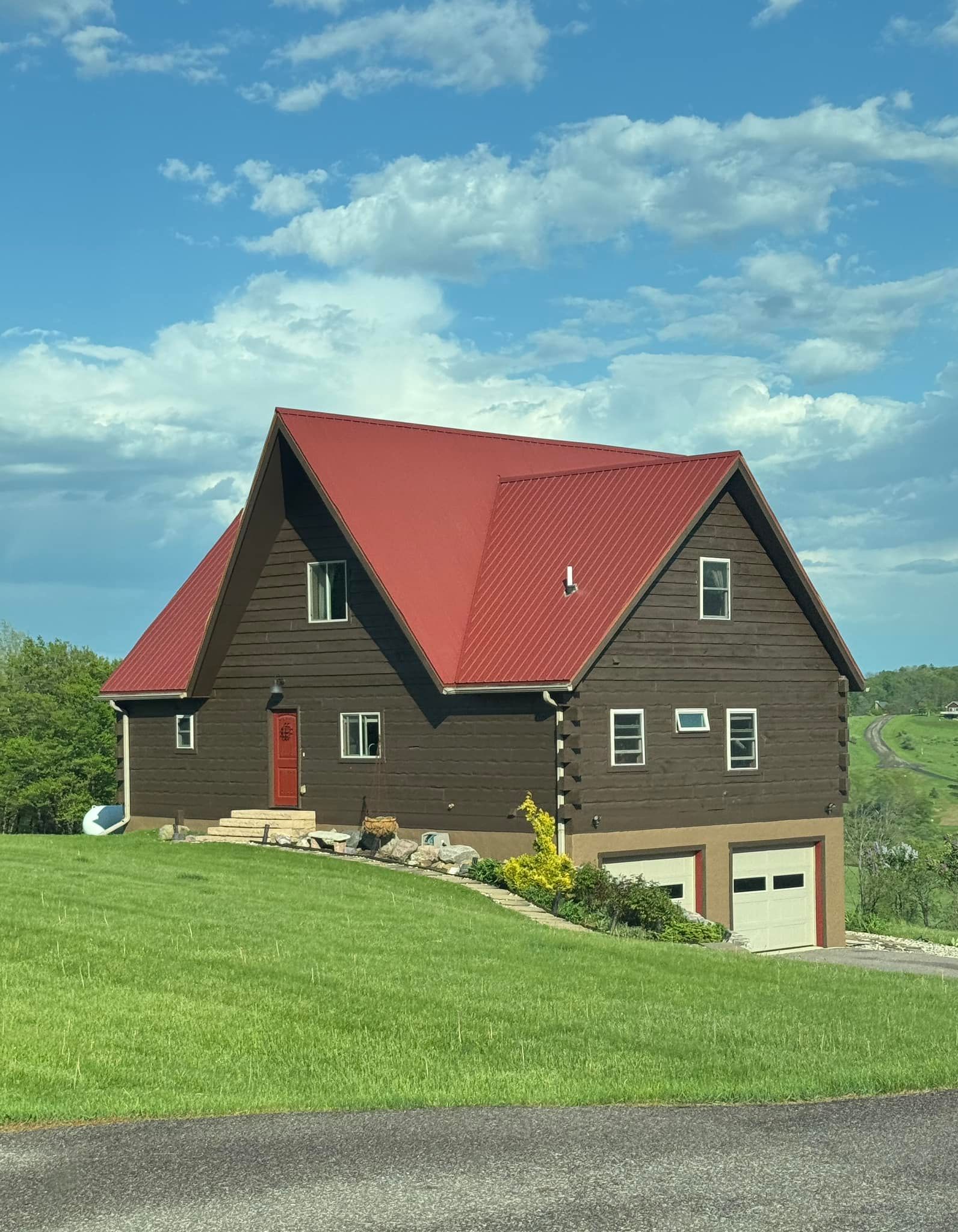 Brown house with a red roof on a grassy hill under a cloudy blue sky. Two garage doors are visible.