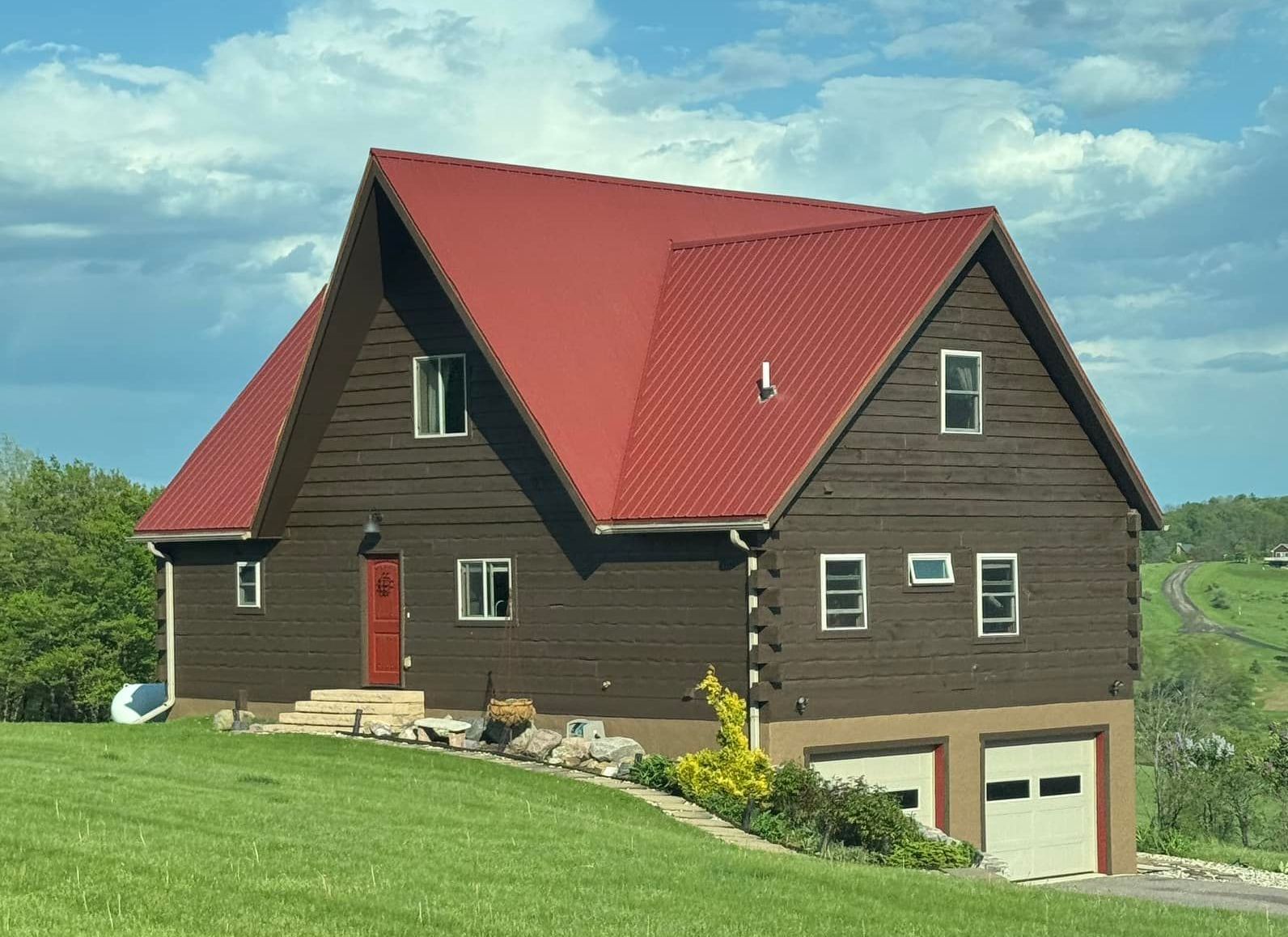 Brown cabin with red roof and two-car garage, set on a grassy hill under a cloudy sky.