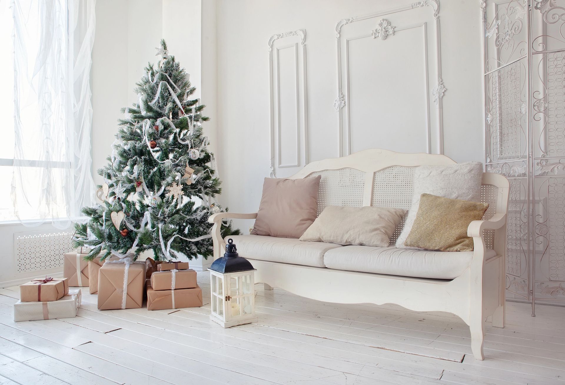 Christmas tree with presents next to a white couch in a bright room.
