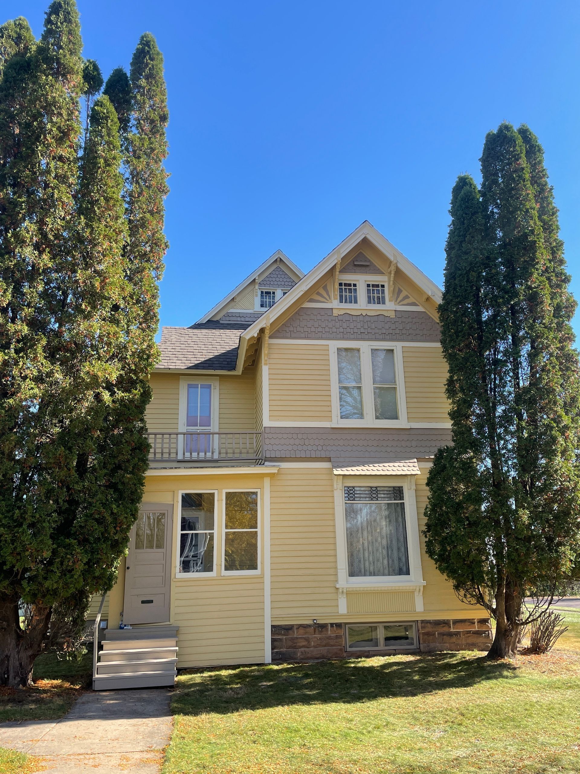 Yellow two-story house with ornate trim, a small balcony, and tall evergreen trees on a sunny day.