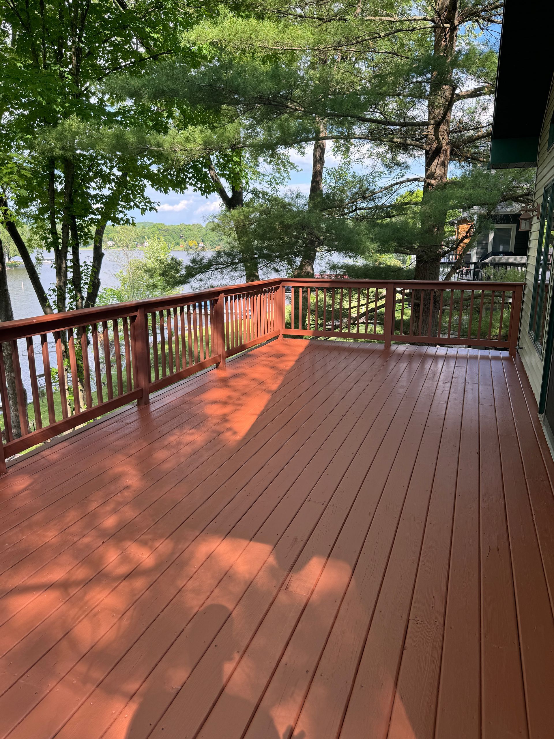 Wooden deck with red-brown stain overlooking a lake. Trees and sky in the background.