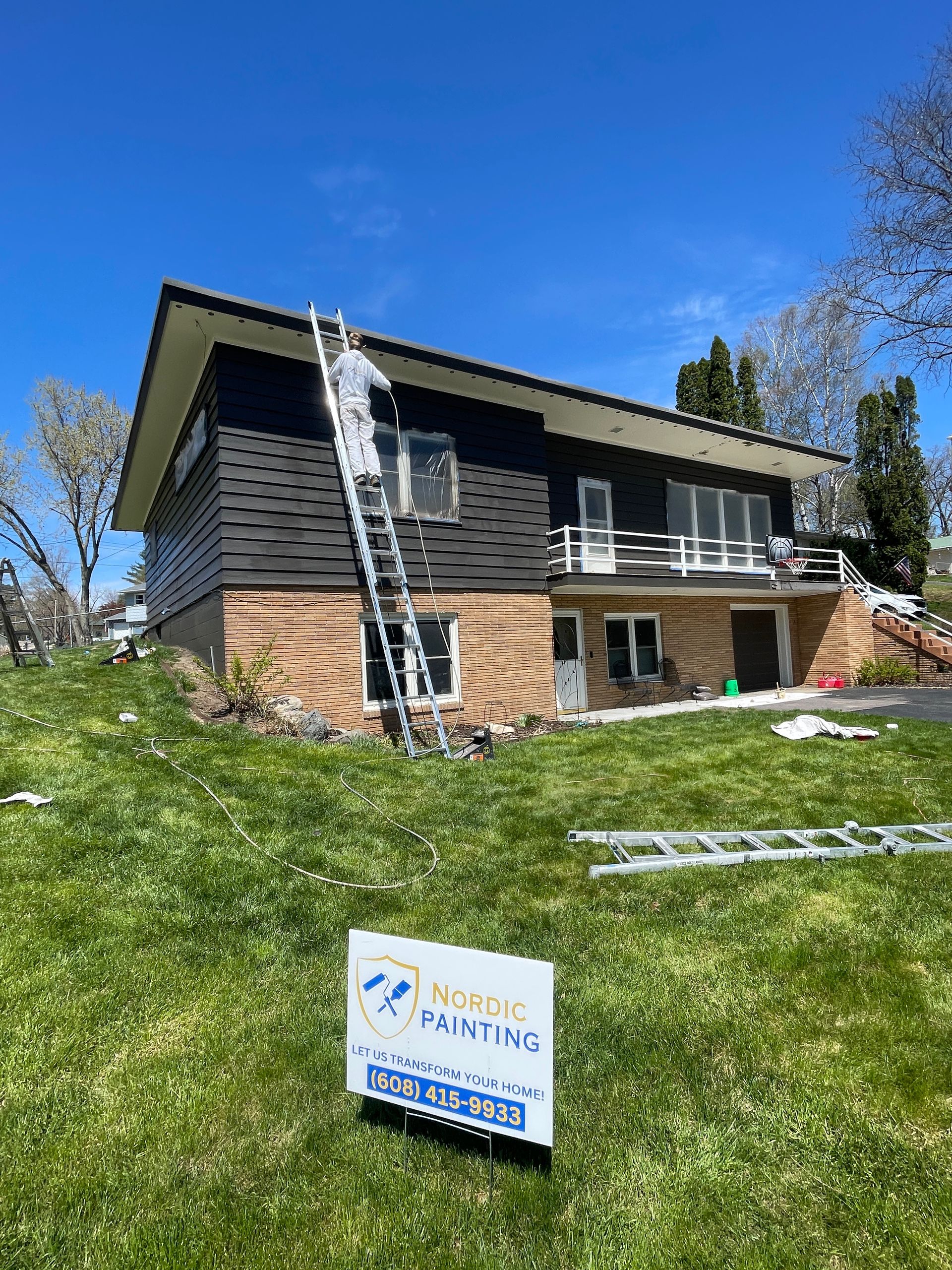 House with black siding, ladder propped against it; signage in the foreground.
