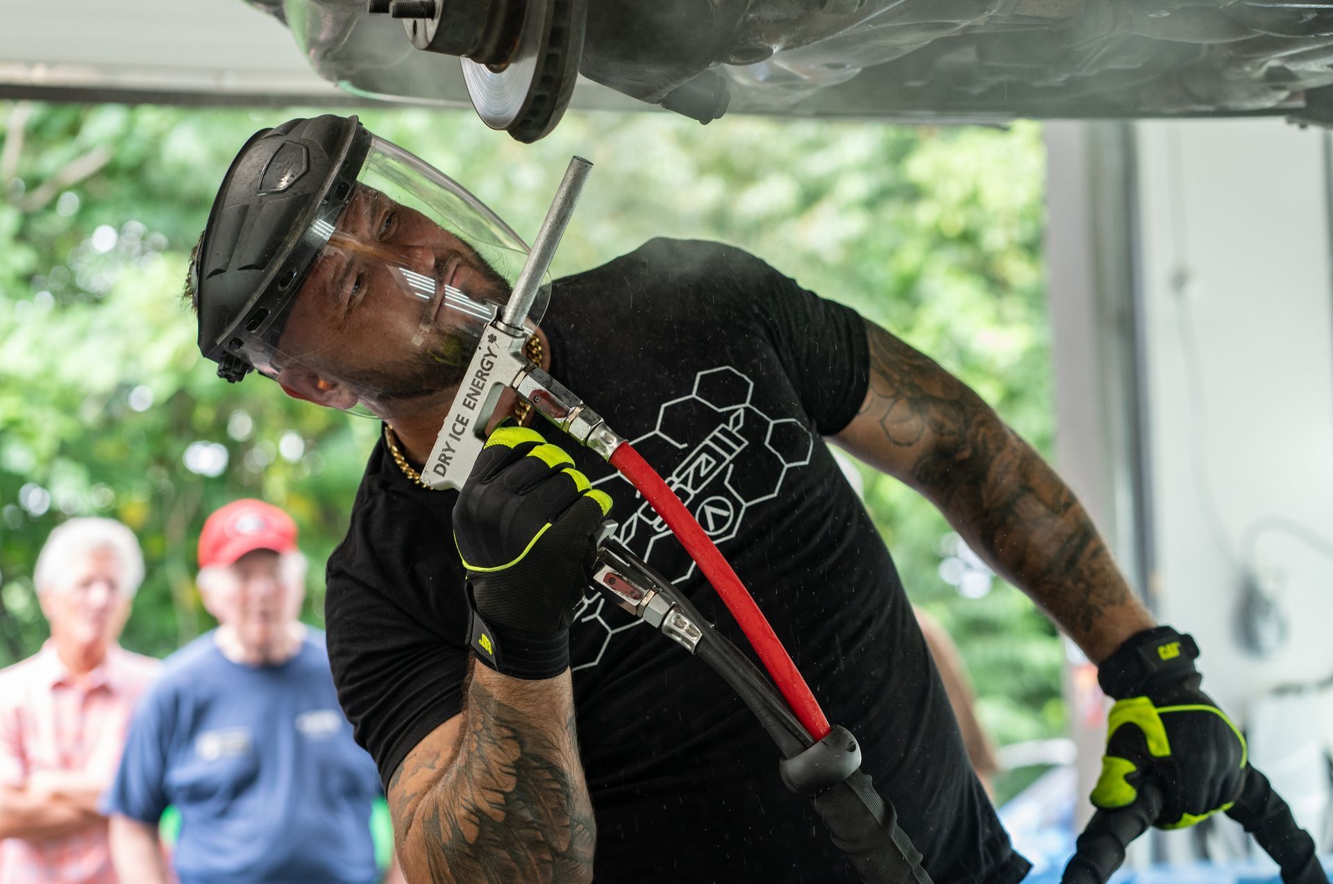 Worker using dry ice blaster under a vehicle, wearing gloves and safety gear.