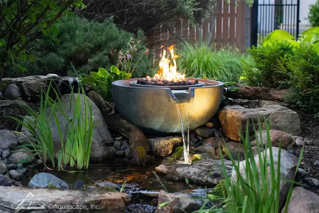 Fire bowl water feature: flame above bowl, water cascading into pond, surrounded by rocks and greenery.