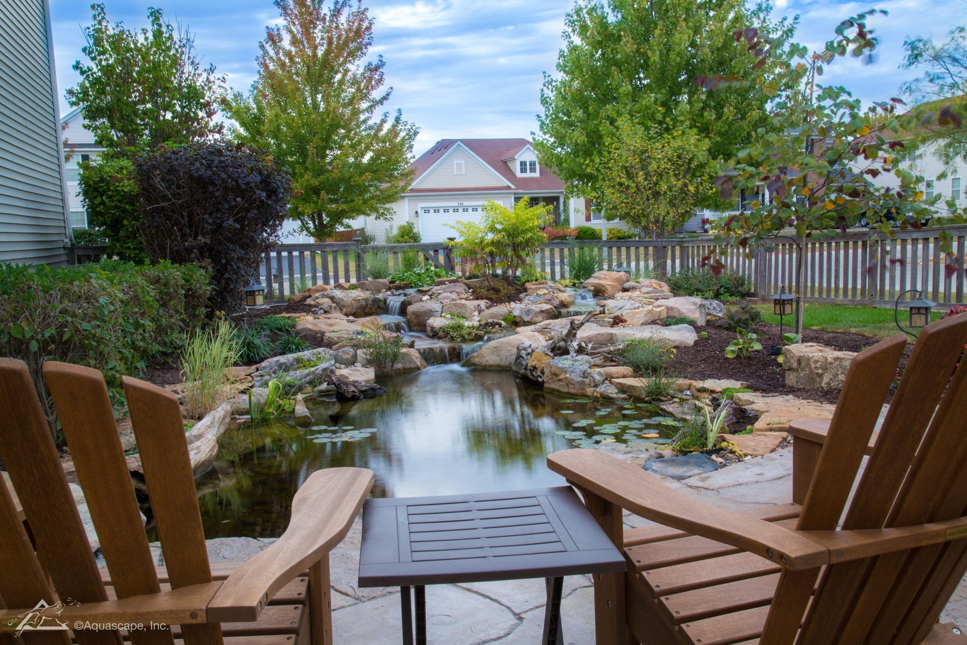 Two wooden chairs face a pond and small waterfall in a backyard.