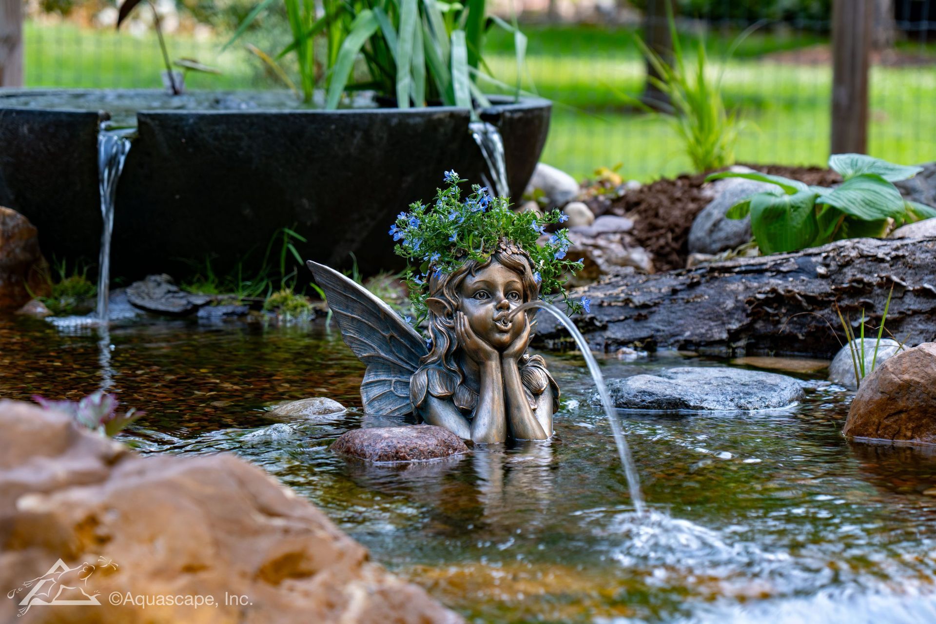 Fairy statue in pond with flowing water, plants, and fountain against a blurred green backdrop.