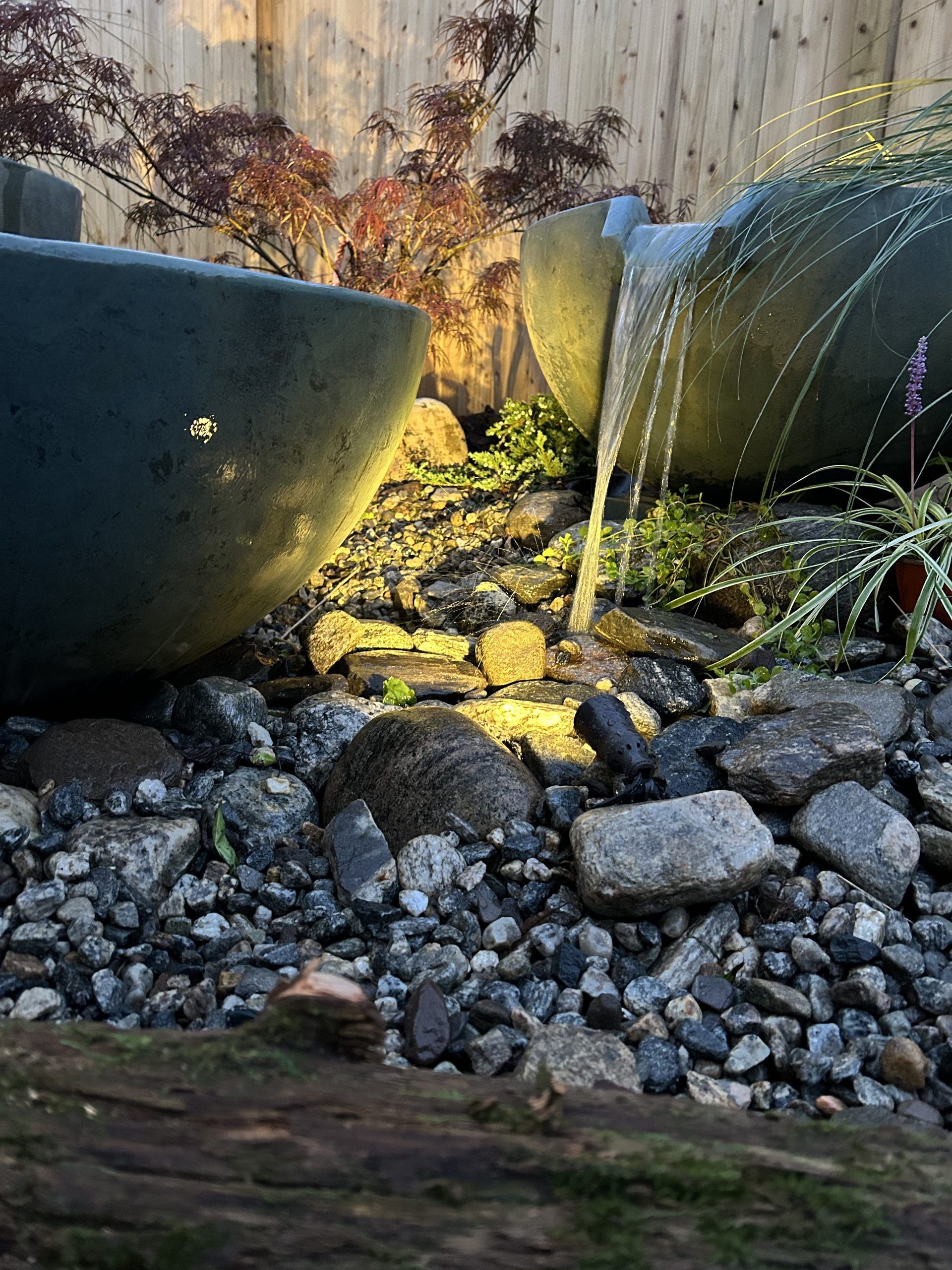 Water feature with two dark bowls, water spilling into rocks, underlit by a warm light; foliage in the background.
