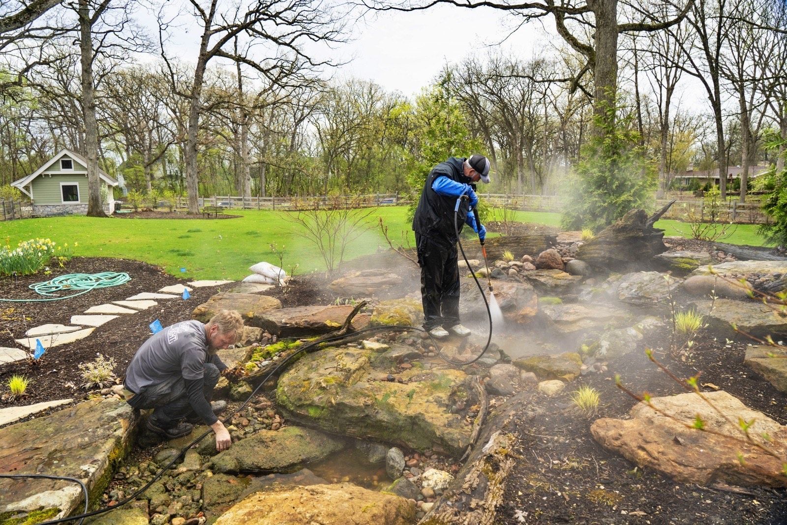 Two people spraying a garden bed with water. One person kneels, the other stands. Green grass and trees in background.