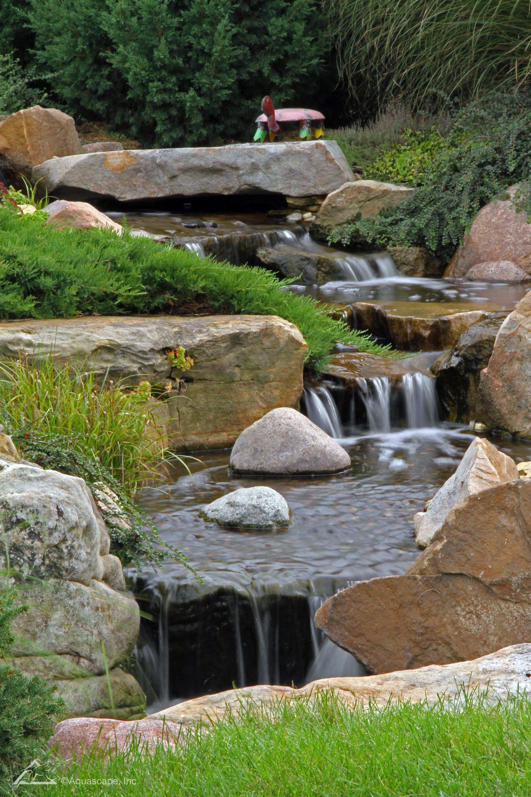 Water cascading down rocks in a naturalistic garden with lush greenery and a small bench in the background.
