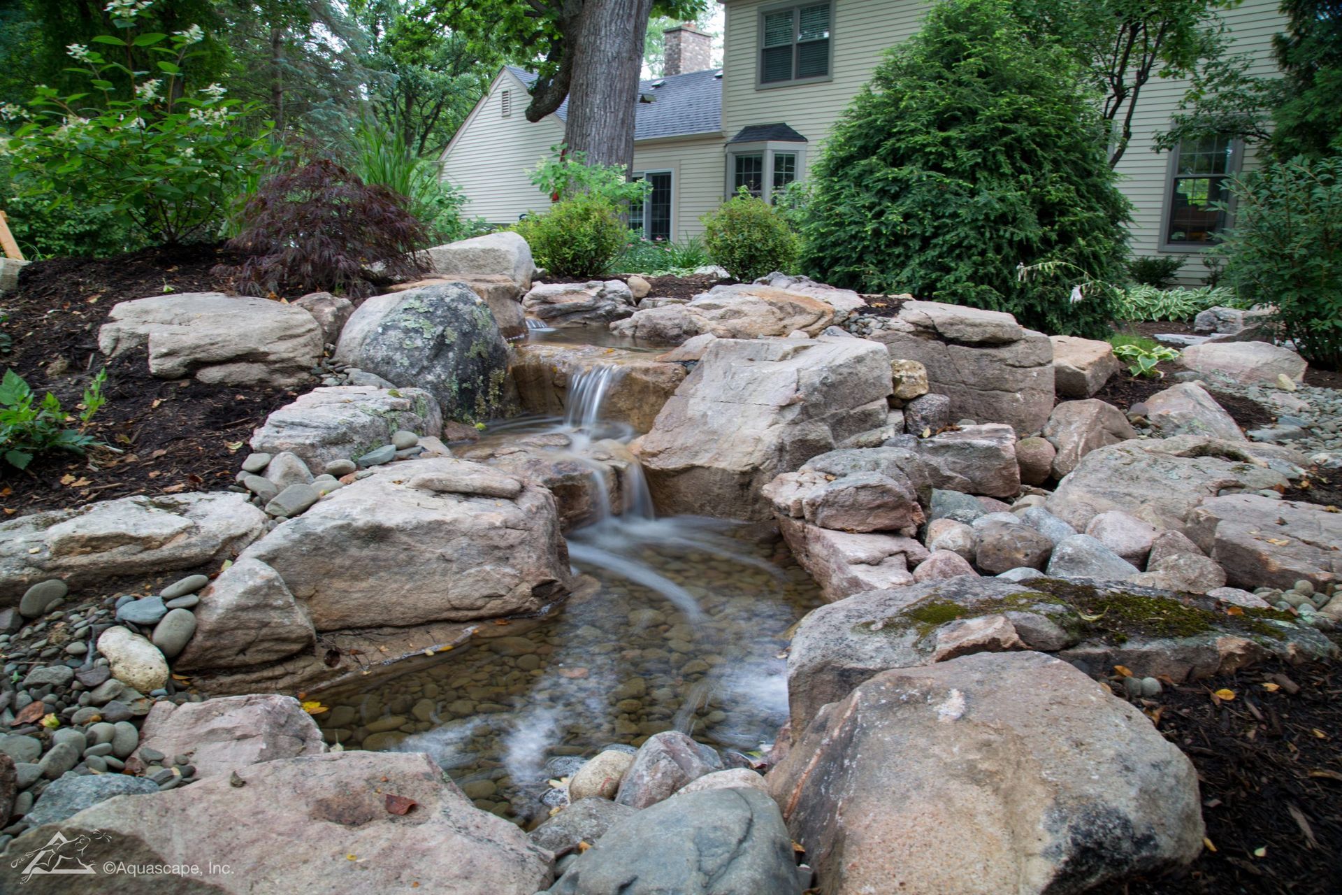 Small waterfall cascading over rocks in a garden; house in background.