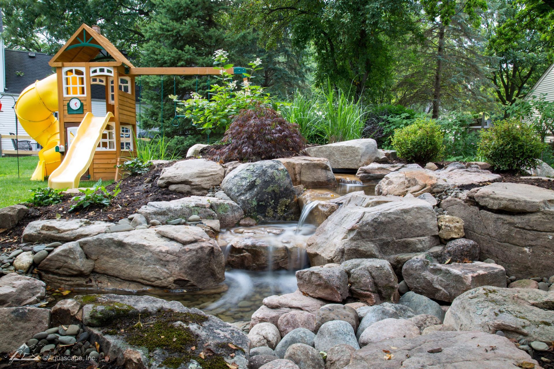 Backyard with a small wooden playhouse, yellow slide, and a rocky waterfall feature.