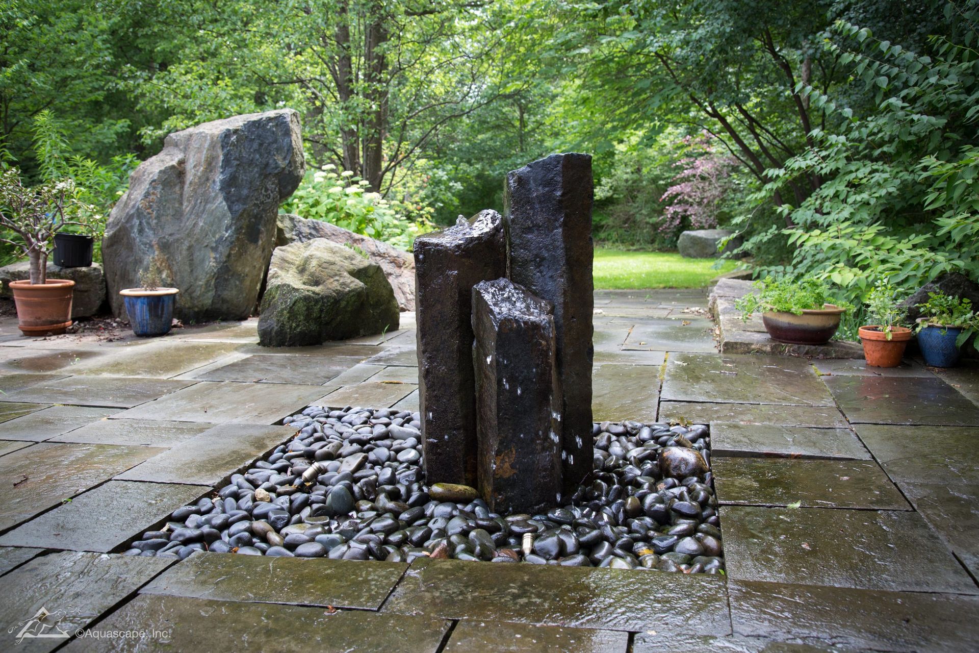 Stone water fountain with dark rocks on a flagstone patio, surrounded by trees and potted plants.