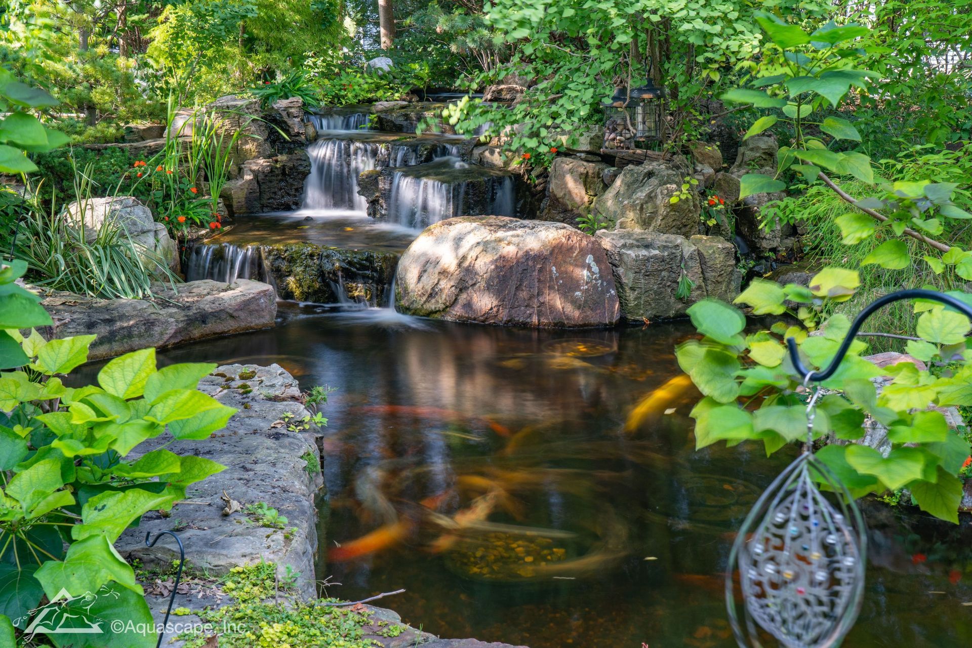 Waterfall cascading into a koi pond surrounded by lush greenery and rocks; a decorative element in the foreground.