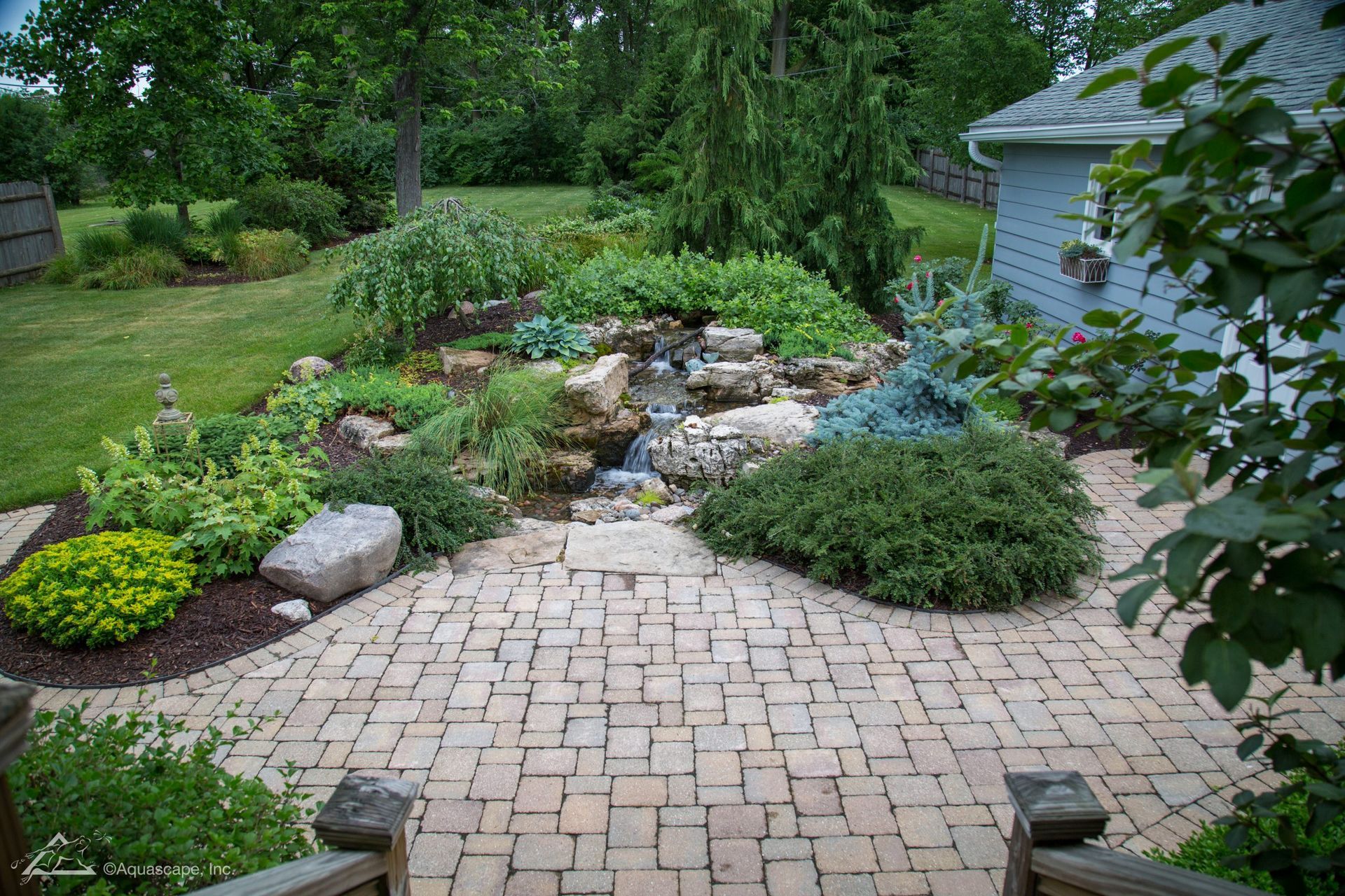 Brick patio leads to a garden with a waterfall, surrounded by lush greenery and a light blue shed.