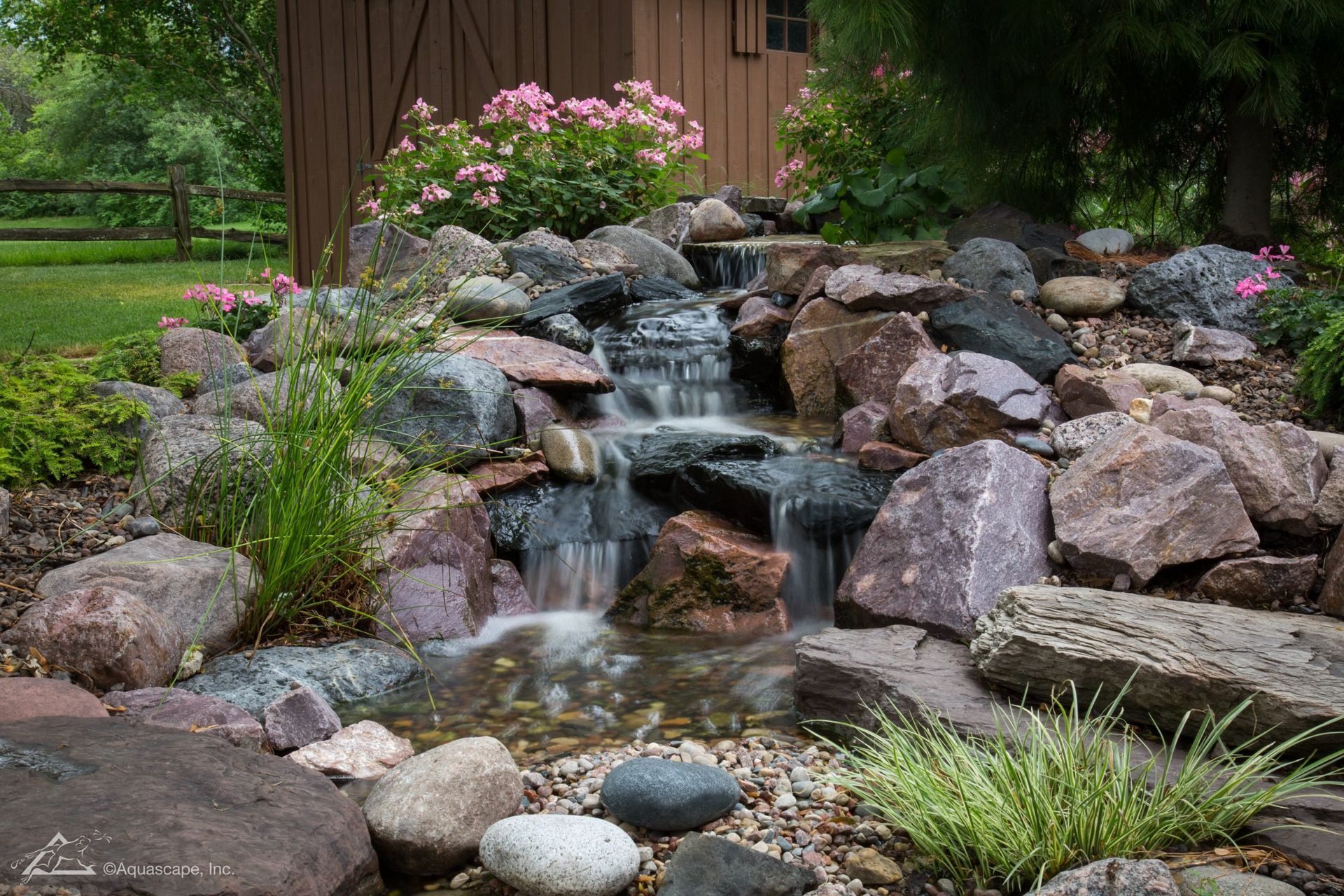Small waterfall cascading over rocks into a pond, surrounded by greenery and flowers, near a brown shed.