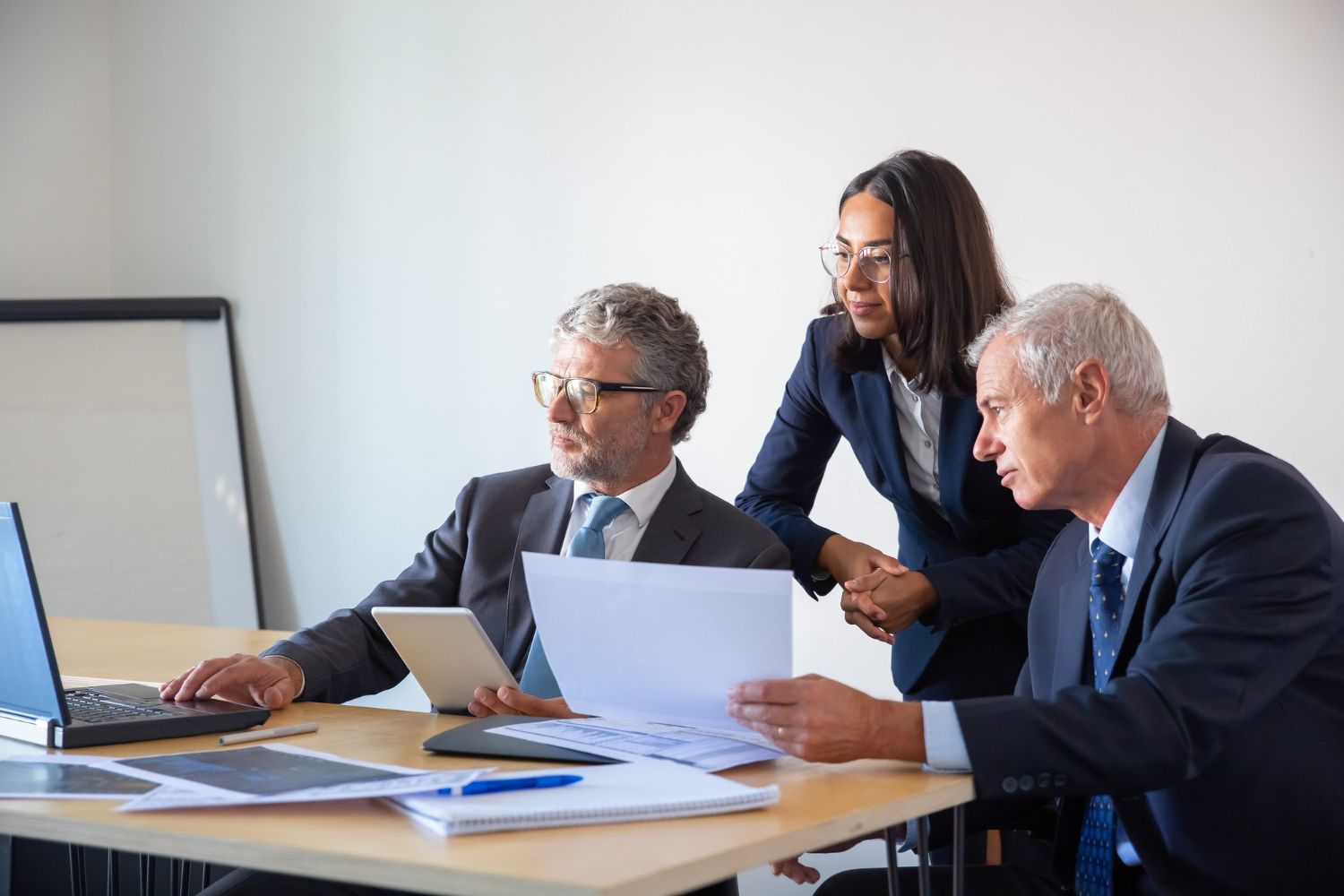 Three coworkers in a meeting reviewing documents at a conference table with a laptop and flip chart.