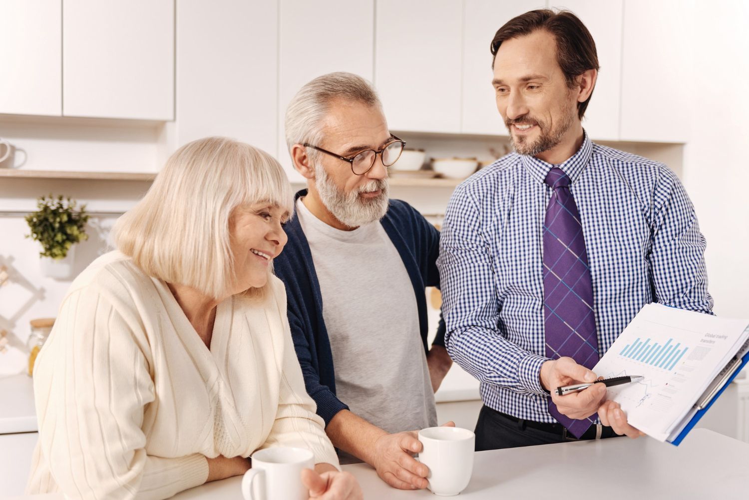 Three people chatting in a bright kitchen, one holding a folder and cup.