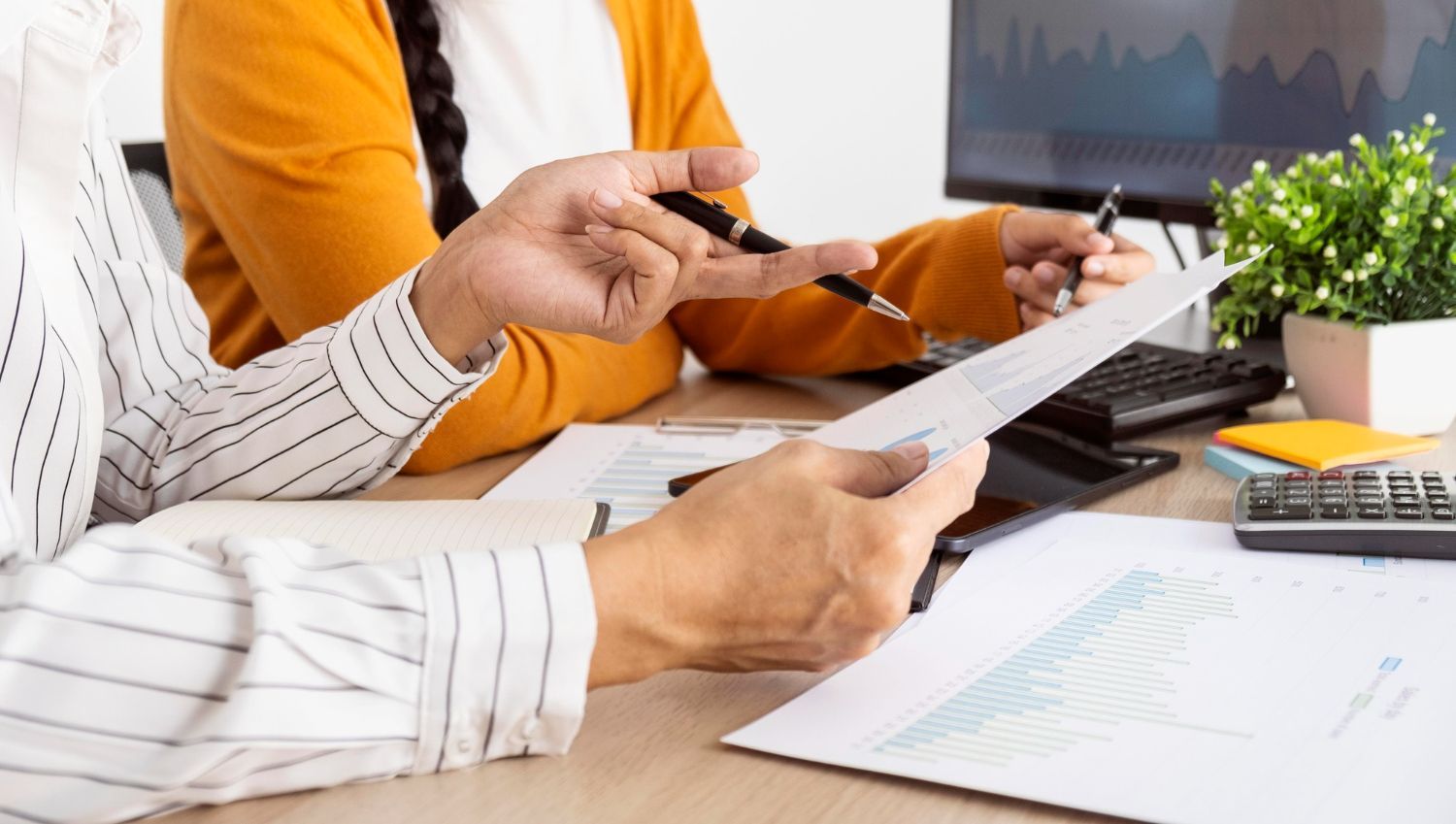 People in business attire reviewing documents and a tablet at a desk with a laptop and plant.