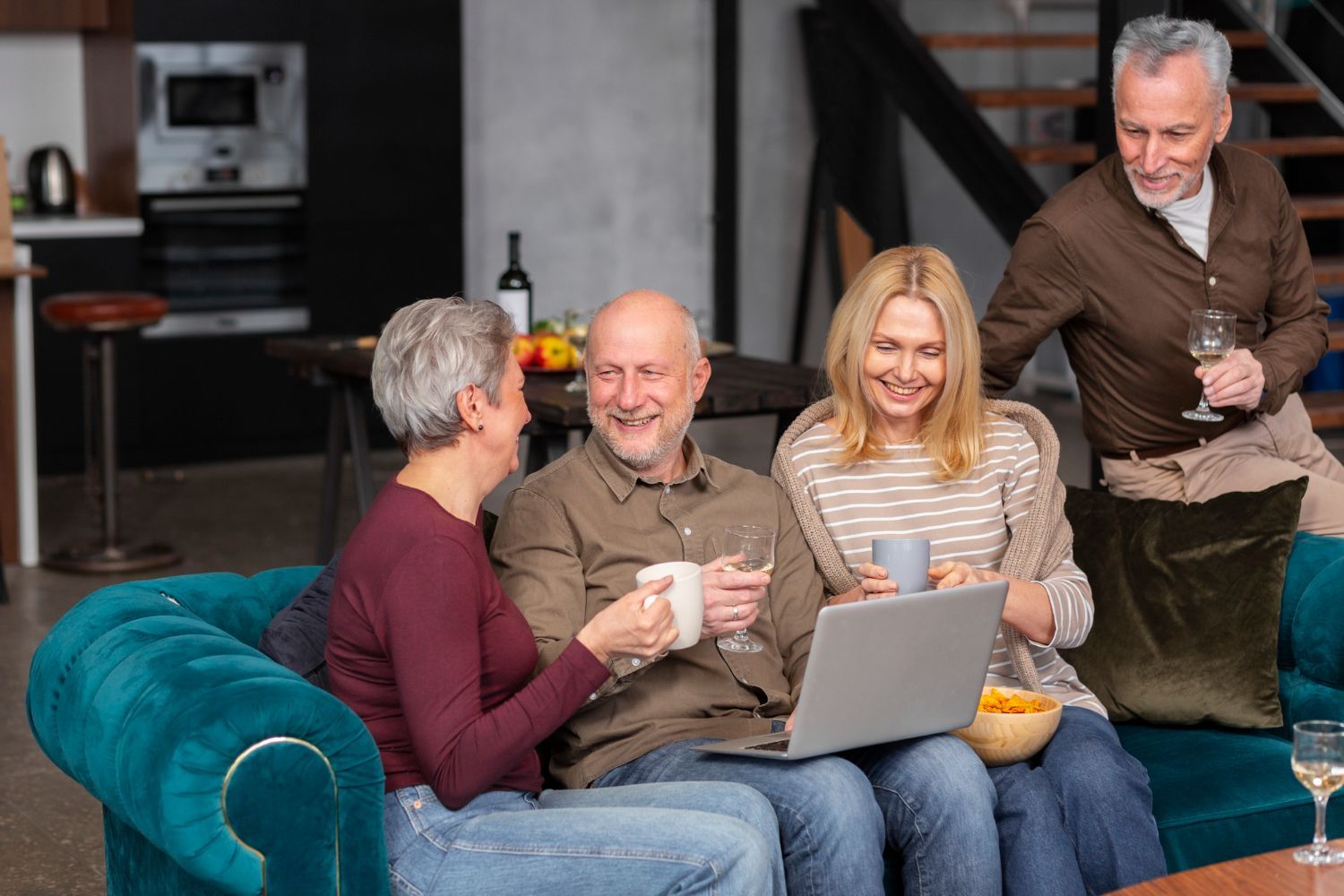 Four people socializing on a teal couch with drinks and a laptop in a modern living room