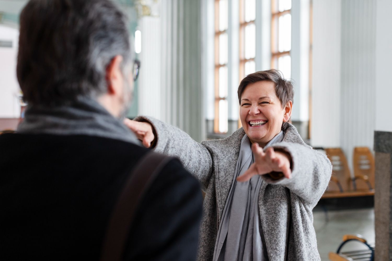 Smiling person in a gray sweater reaching out to greet someone indoors