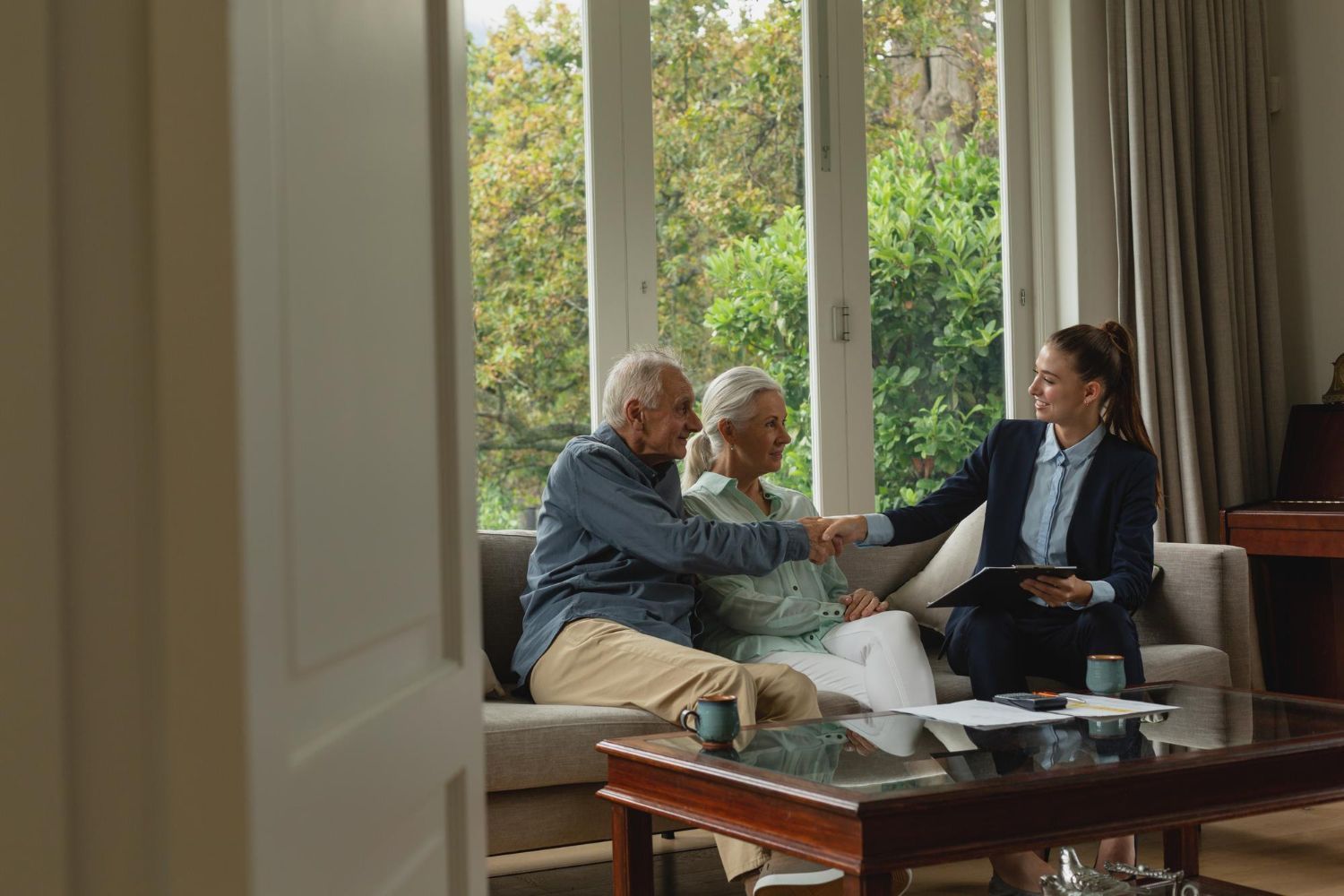 Three people in a living room shake hands during a discussion by a window.