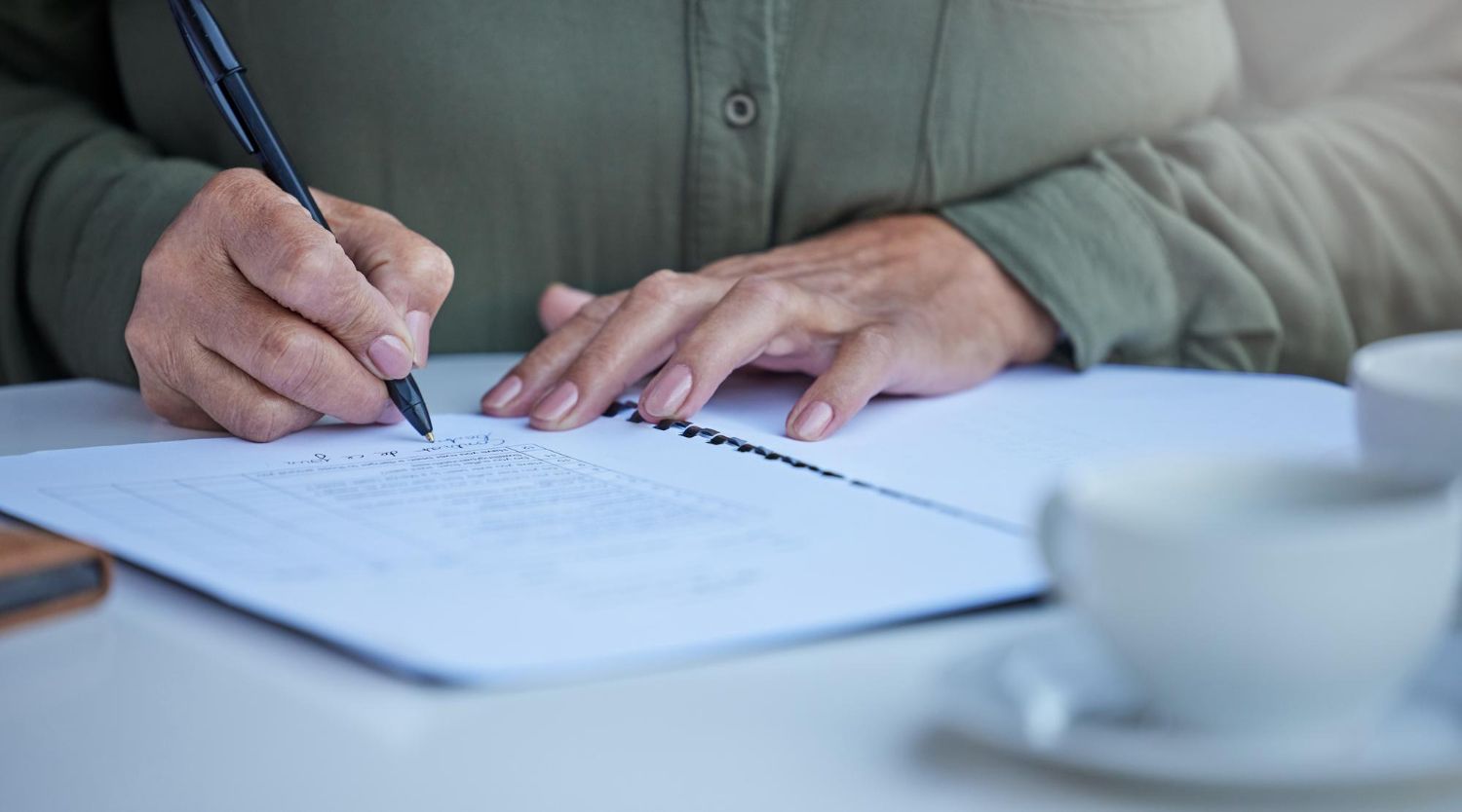 Hands writing with a pen on a document at a table beside a coffee cup