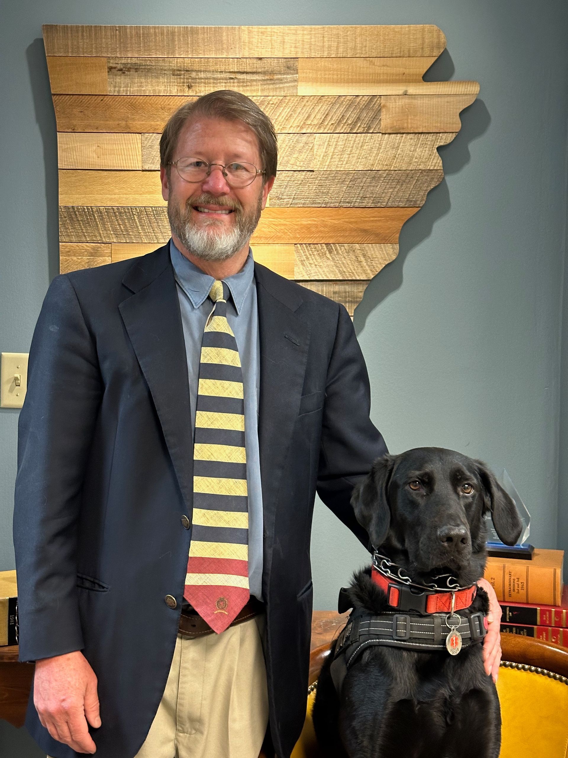 Man in a suit standing beside a black guide dog in a harness, indoors against a wall map.