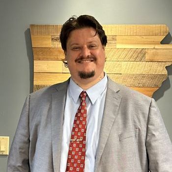 Smiling man in a gray suit and red tie standing before a wooden wall art sign