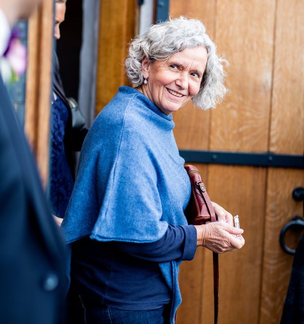 Smiling person in a blue sweater standing by a wooden door, holding a brown bag.