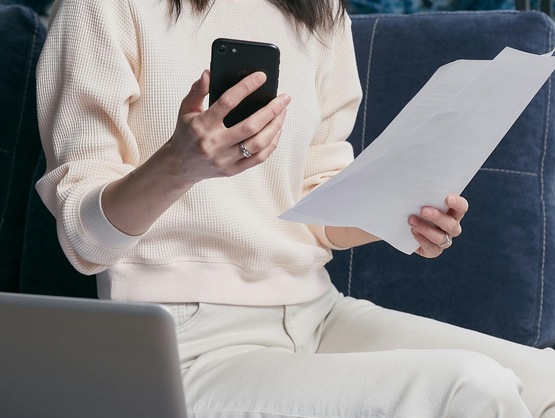 Person holding a phone and reading a document while seated on a blue couch with a laptop nearby