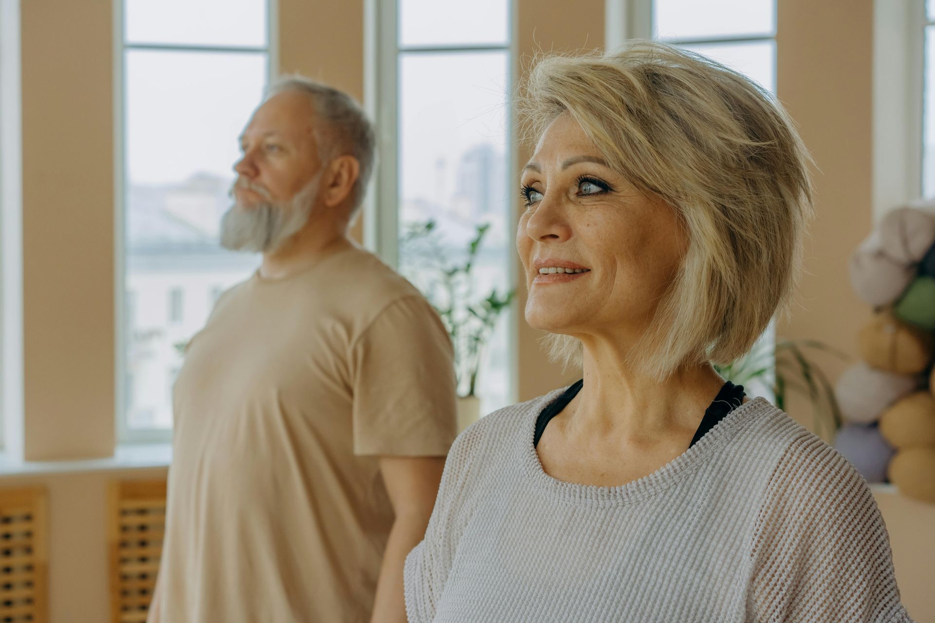 Two people standing in a bright room, one smiling in the foreground and one near a window in the background