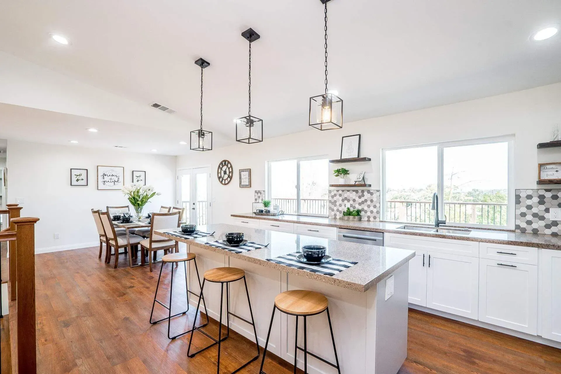 Bright kitchen with island and bar stools, leading to dining area. Wooden floors, white cabinets, three pendant lights.