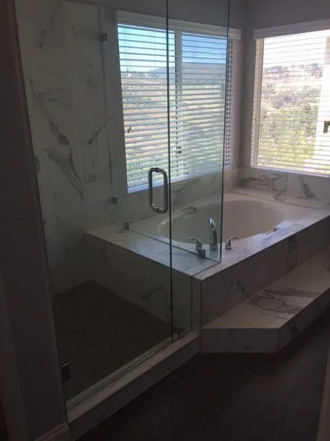 Bathroom with a marble-tiled bathtub and shower, glass enclosure, and window with blinds.