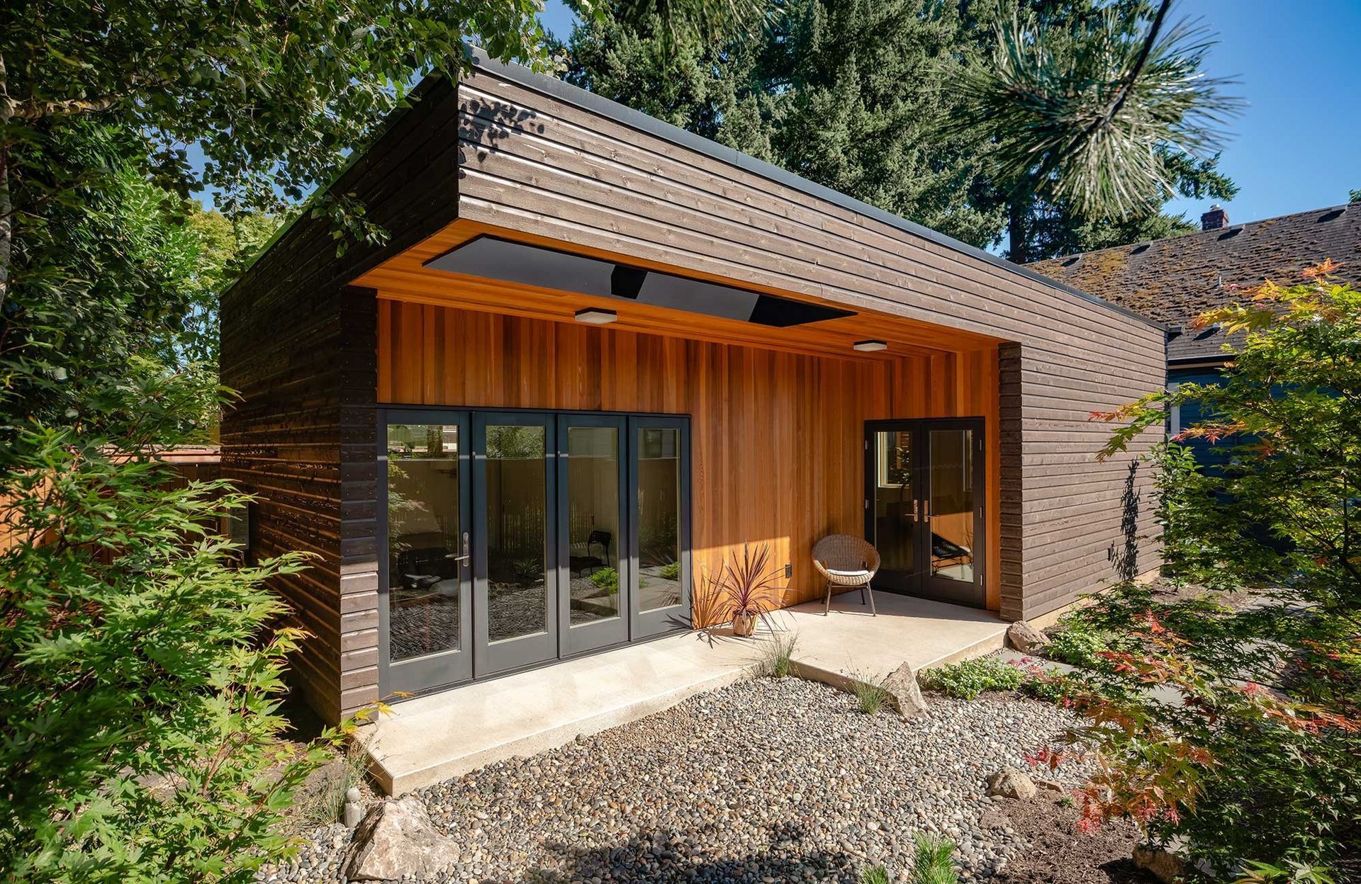 Modern wood house with sloped roof, large windows, and gravel yard surrounded by trees