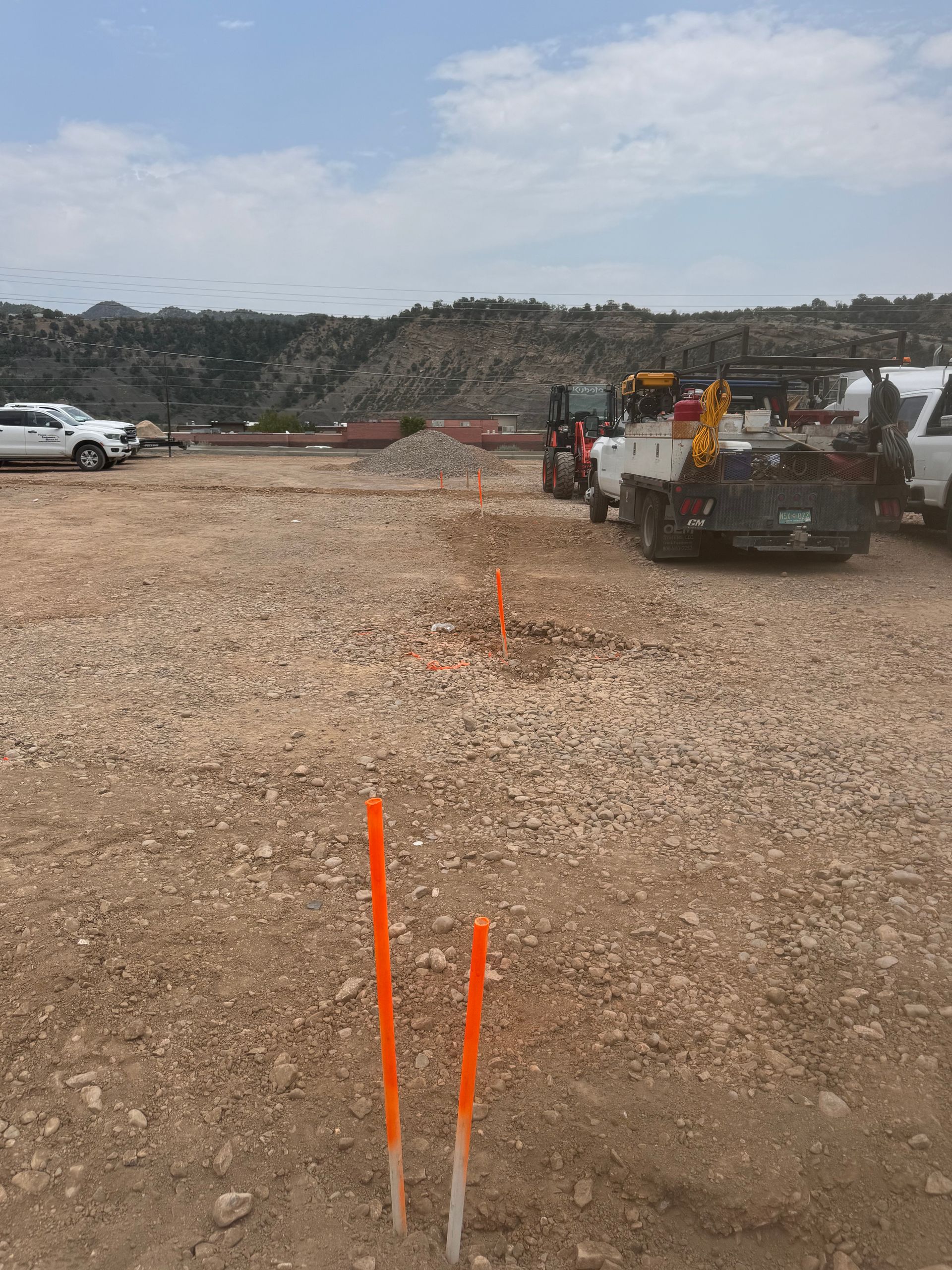 Orange stakes mark a path in a gravel lot, with trucks and a large pile of debris in the background.