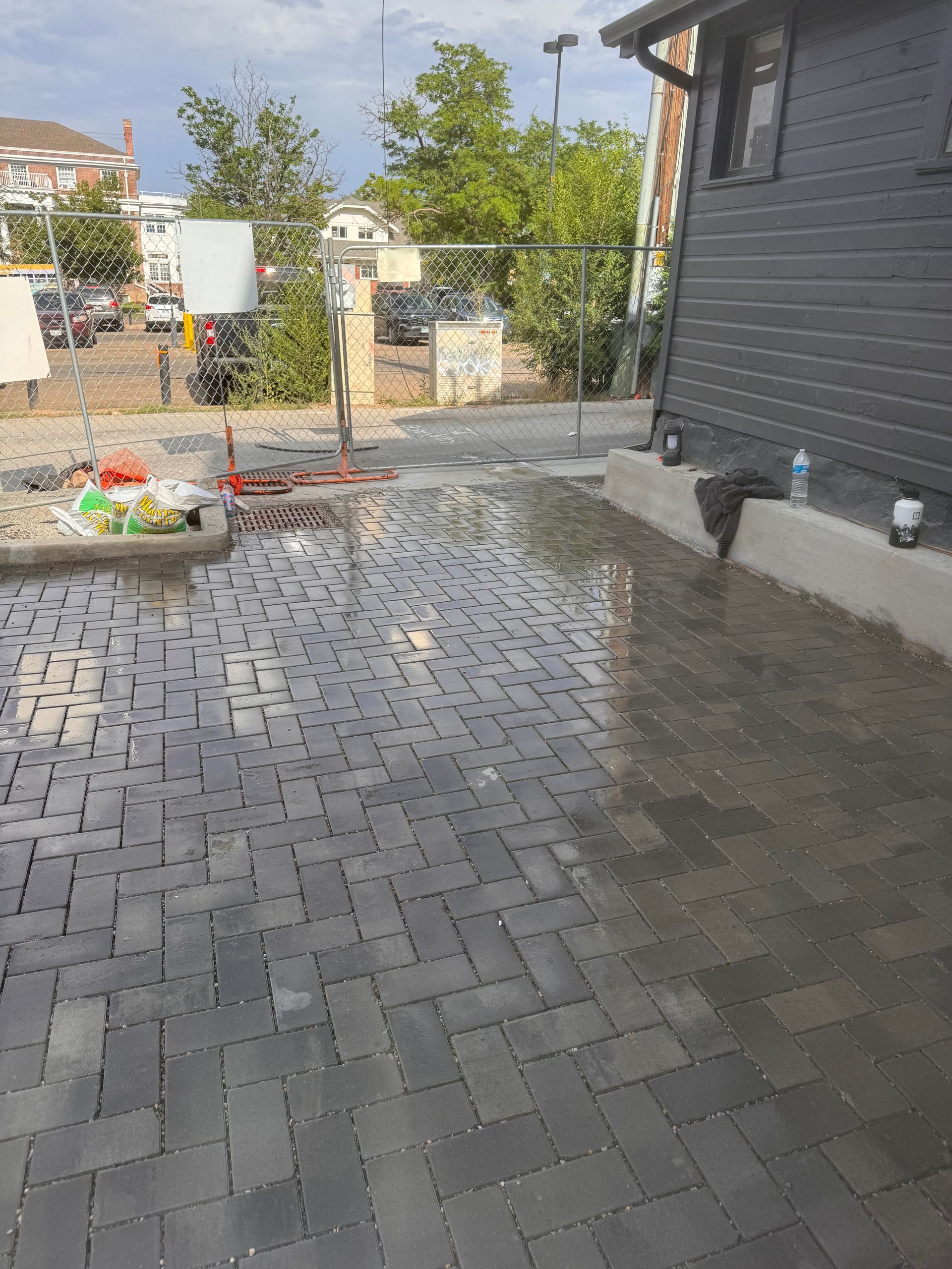 Wet, dark gray brick paved patio next to a dark gray wooden building, with a few construction materials visible.